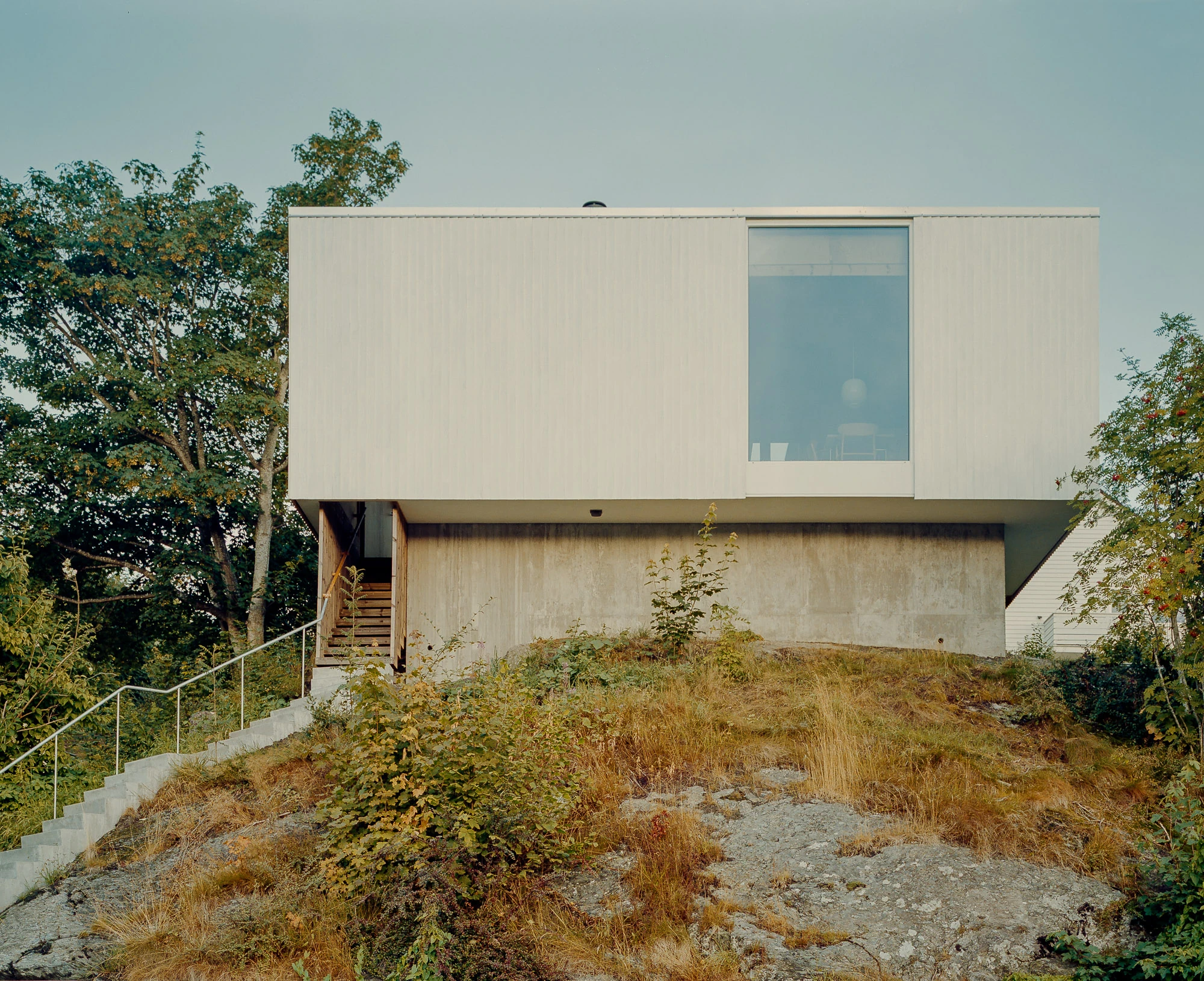 Ersland & Skeie House by Knut Hjeltnes Architects, photographed by Hugo Lütcherath — The upper floor hovers above a concrete wall and bare bedrock, its white cladding blending with the surrounding white buildings.