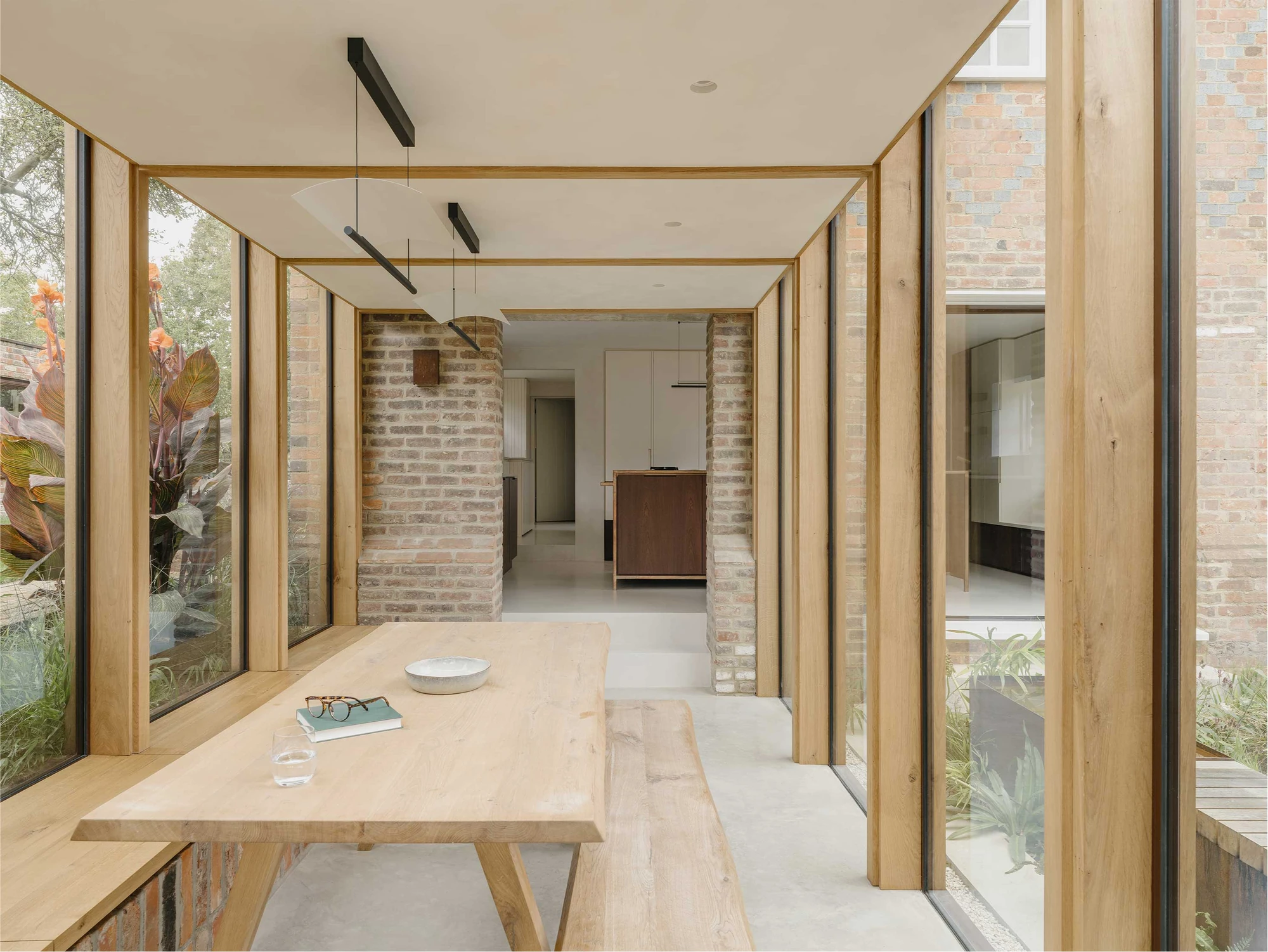 Muttram Veedu by Will Gamble Architects, photographed by James Retief — An oak dining table and bench sit in the glazed extension, brick piers framing the route back to the kitchen and its polished concrete floor.