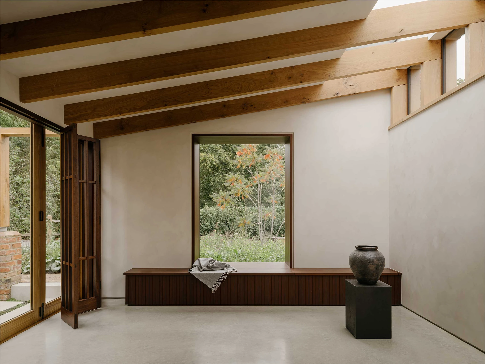 Muttram Veedu by Will Gamble Architects, photographed by James Retief — A deep window reveal frames a young tree, oak beams overhead and a clerestory strip lifting the roof from the wall.