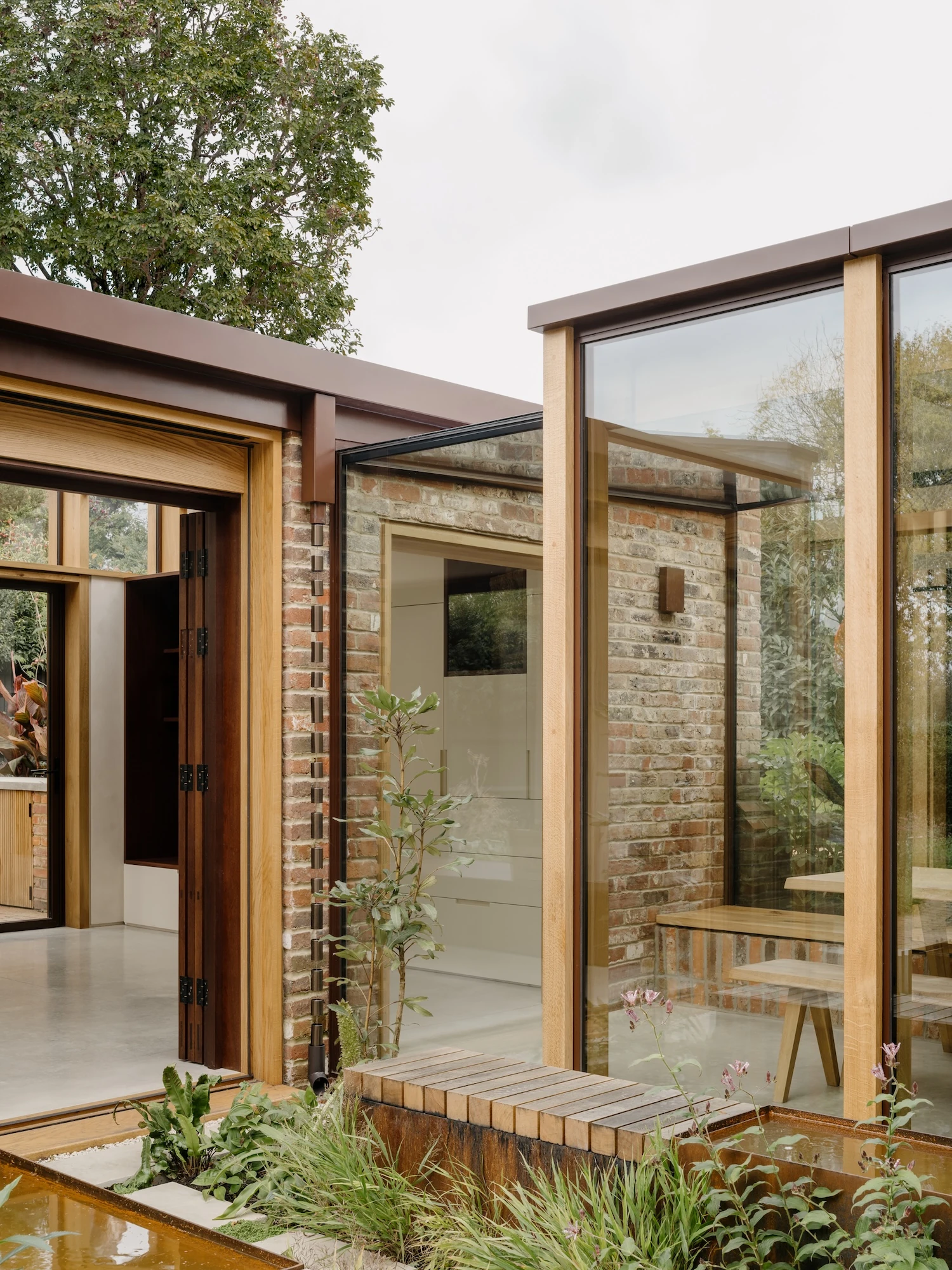 Muttram Veedu by Will Gamble Architects, photographed by James Retief — The glazed link stands in the courtyard behind tall planting, its timber frame and dark parapet set against reclaimed brick.