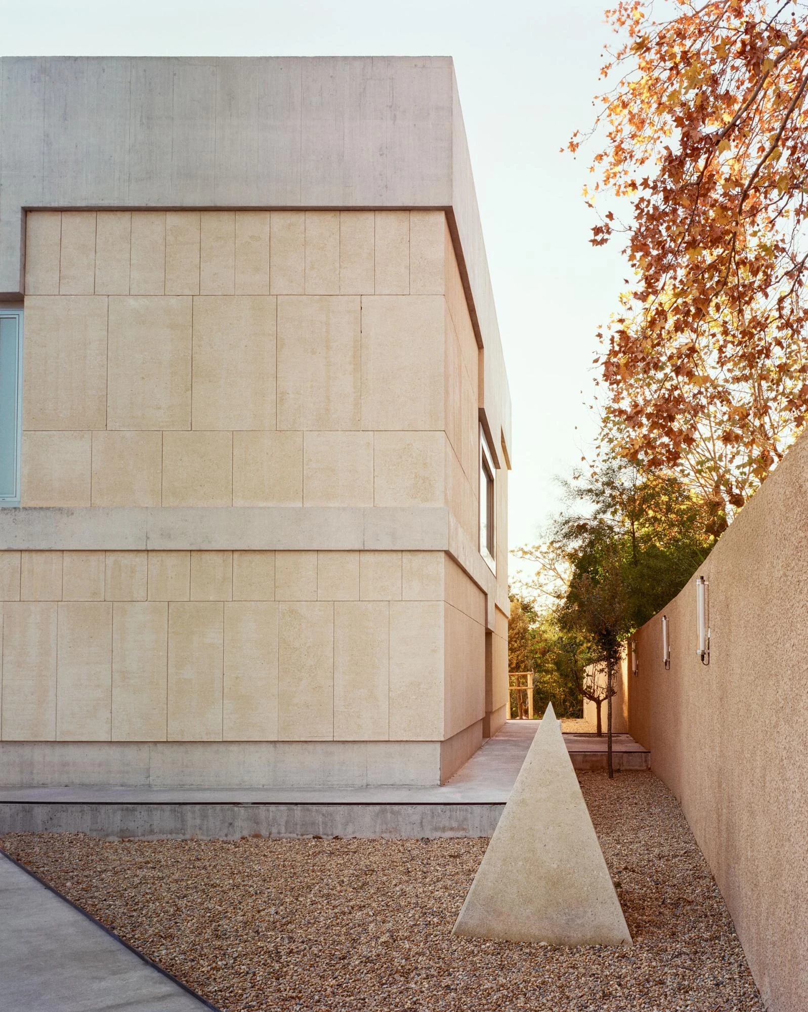 Cultural and Community Center, Coudoux by atelier EGR, photographed by Giaime Meloni — A stone pyramid stands on the ground beside the main building's stone volume and raw concrete base.
