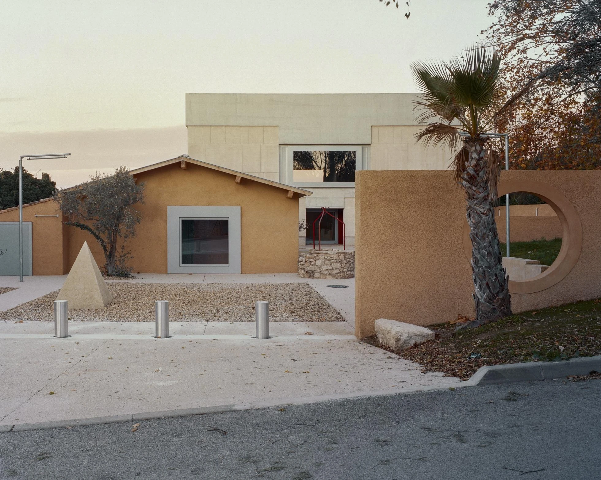 Cultural and Community Center, Coudoux by atelier EGR, photographed by Giaime Meloni — At dusk the sheepfold, the main building, the old well and a stone pyramid line the public square.