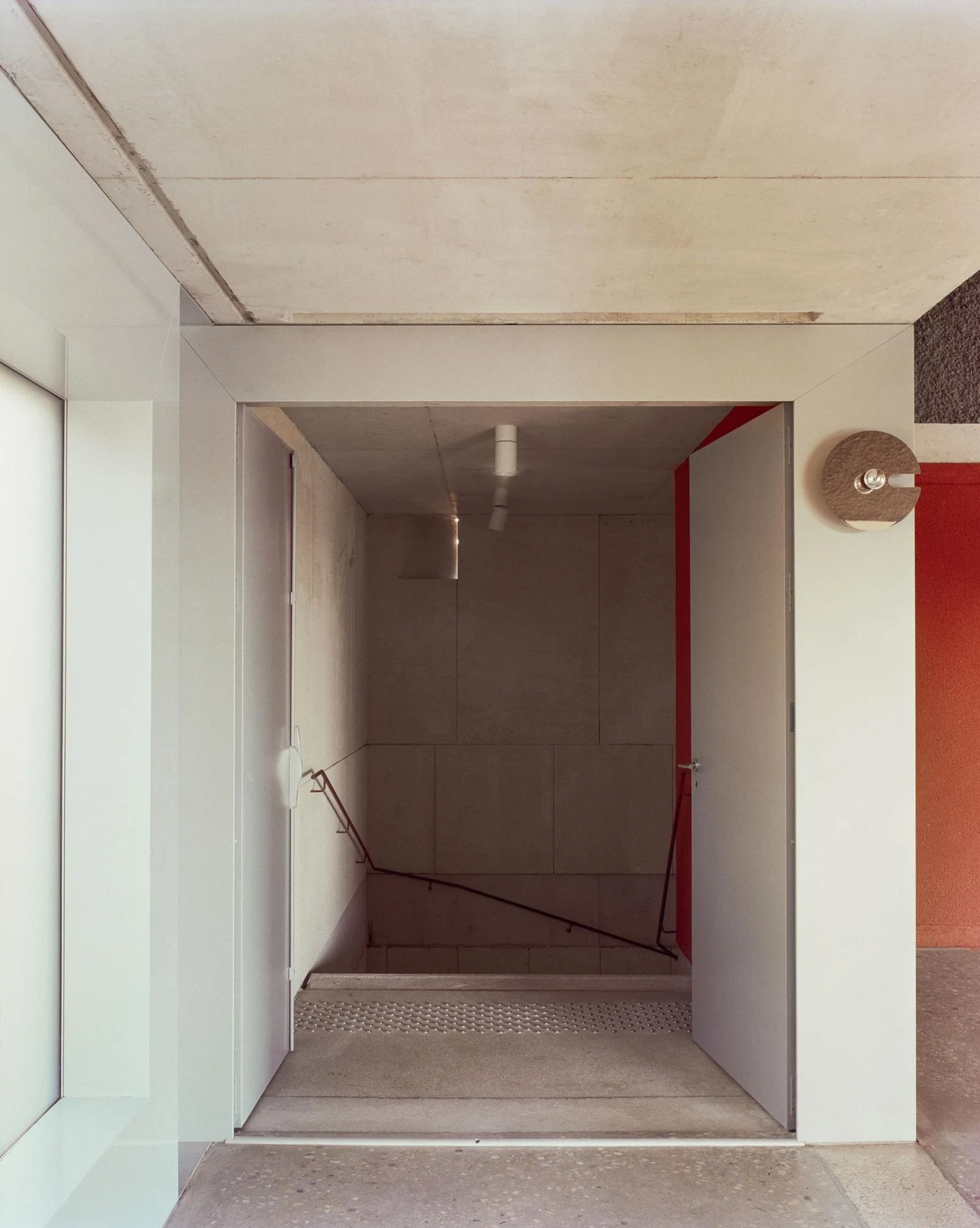 Cultural and Community Center, Coudoux by atelier EGR, photographed by Giaime Meloni — An interior threshold opens onto a concrete stair descending between raw concrete walls, red earth plaster to one side.