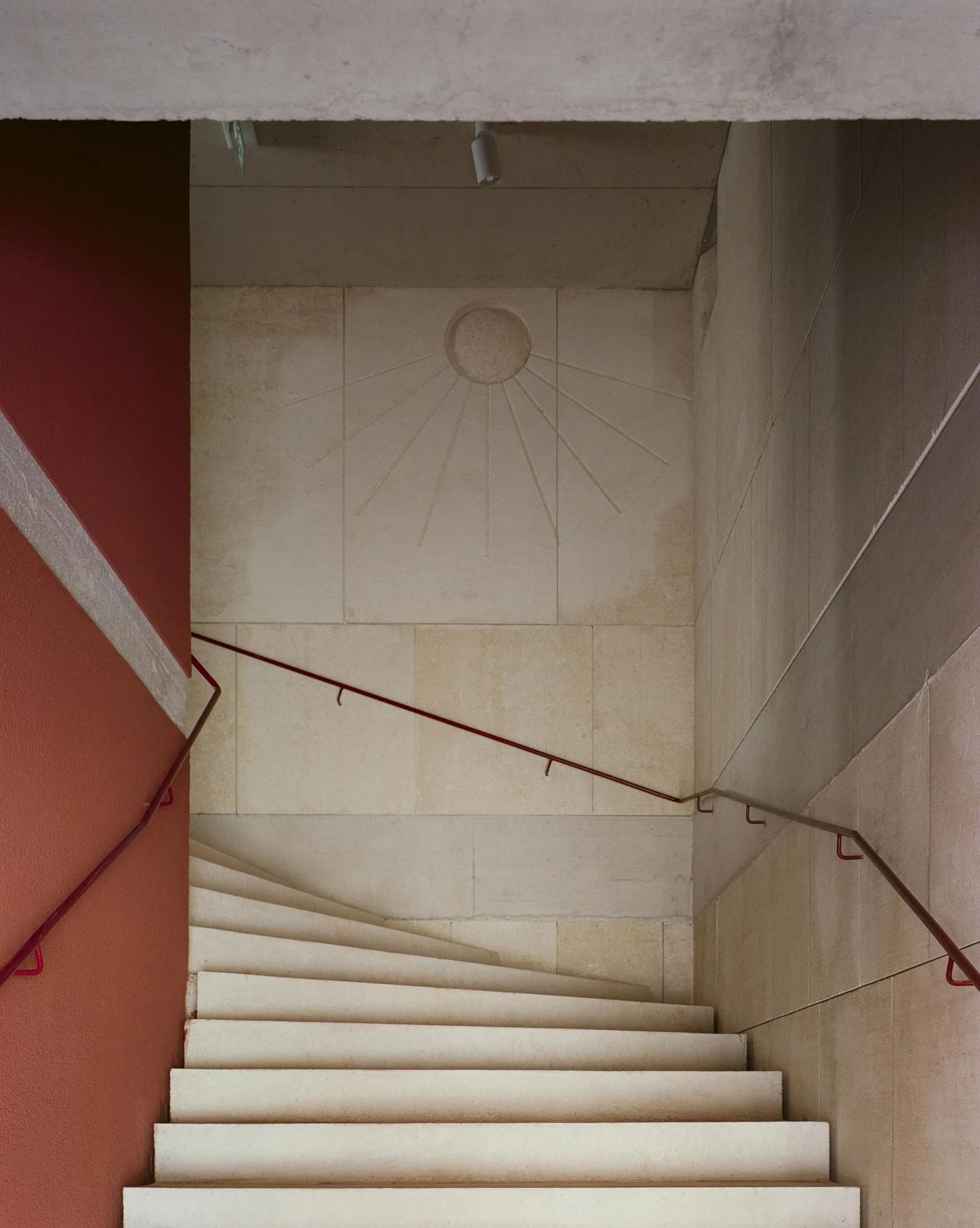 Cultural and Community Center, Coudoux by atelier EGR, photographed by Giaime Meloni — A concrete stair turns past a wall where a sun is engraved into the stone, red earth plaster alongside.