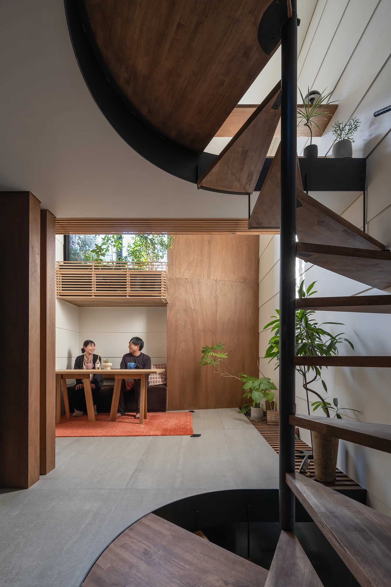 House in Kagurazaka by Takumi Wakui + Riho Wakui Architects — A multi-storey void opens through the timber-lined interior, framed greenery visible beyond as the couple sit at a table on the floor below.