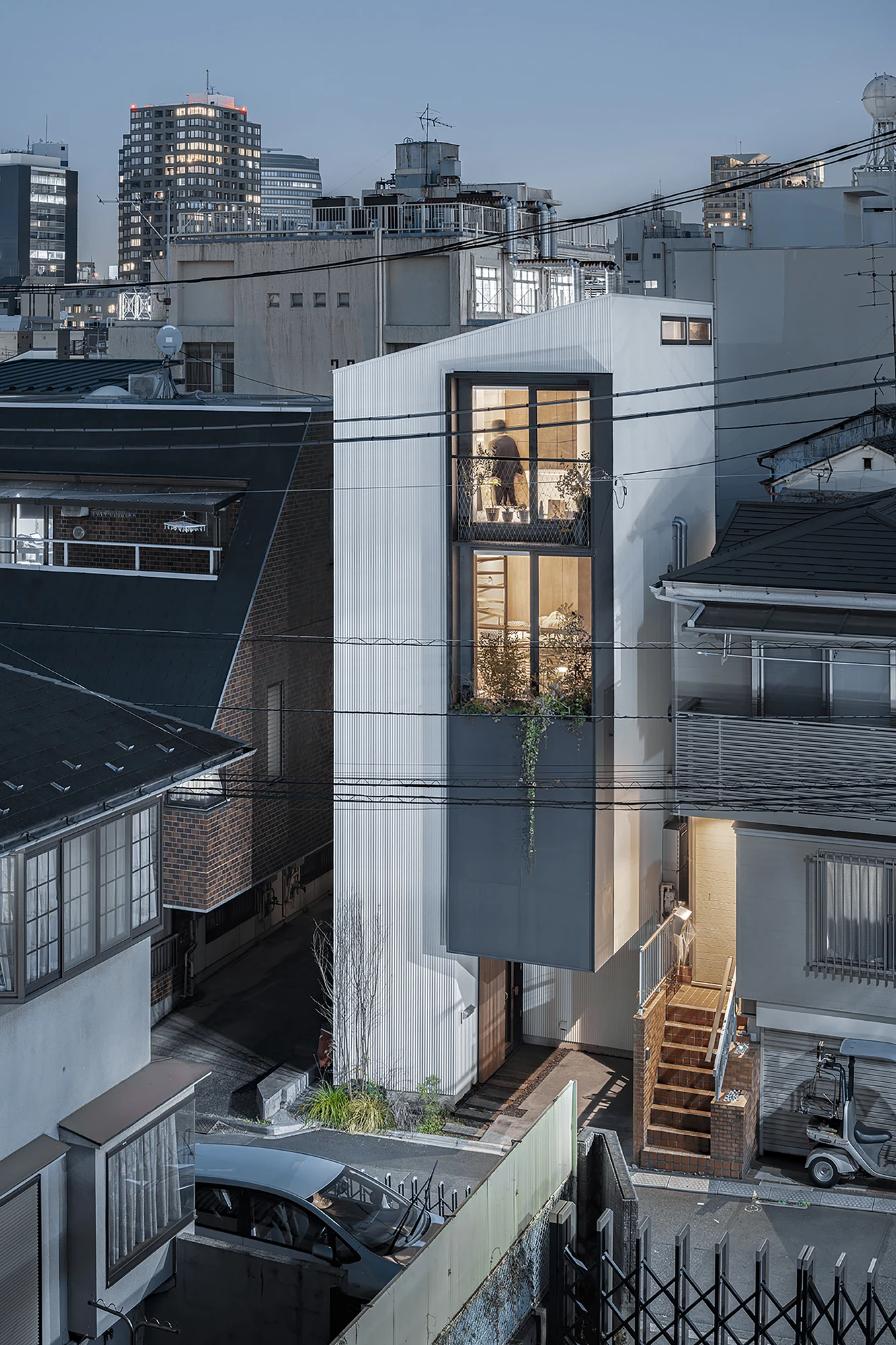 House in Kagurazaka by Takumi Wakui + Riho Wakui Architects — At dusk the lit interior glows through the street-facing framed windows, the four-storey timber house standing amid the dense district.