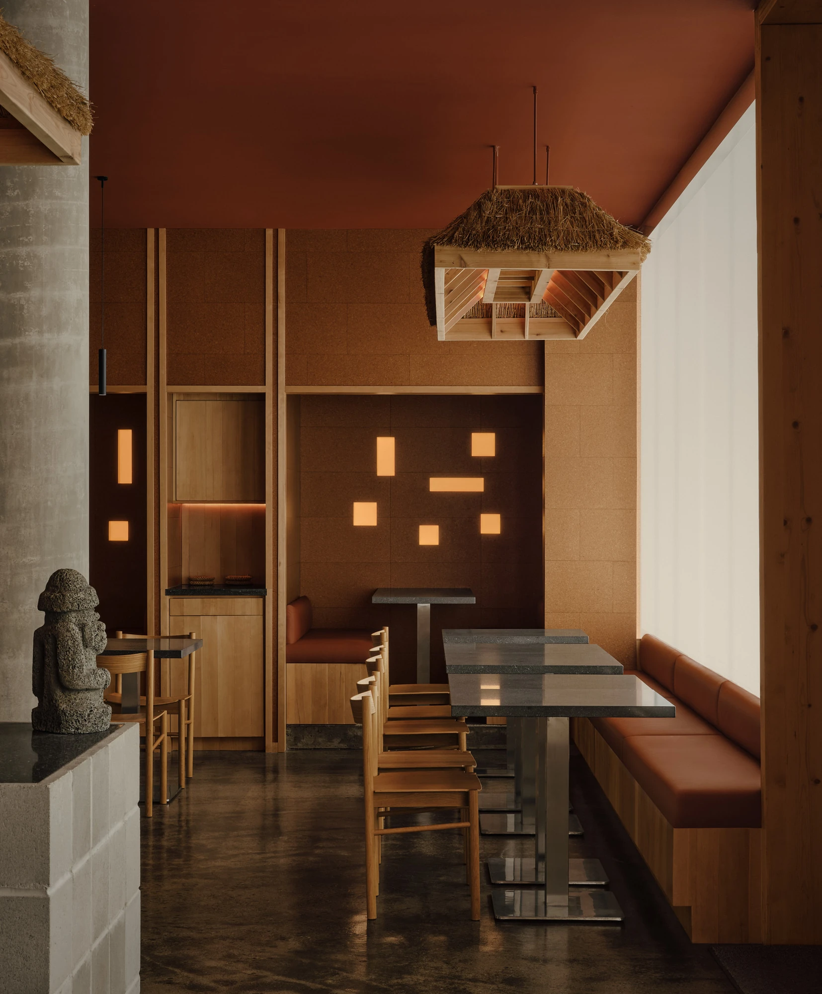 Jeju by &Daughters, photographed by Alex Lesage — Banquette seating along the cork wall, framed by timber ceiling beams and the underside of a hand-thatched fixture.