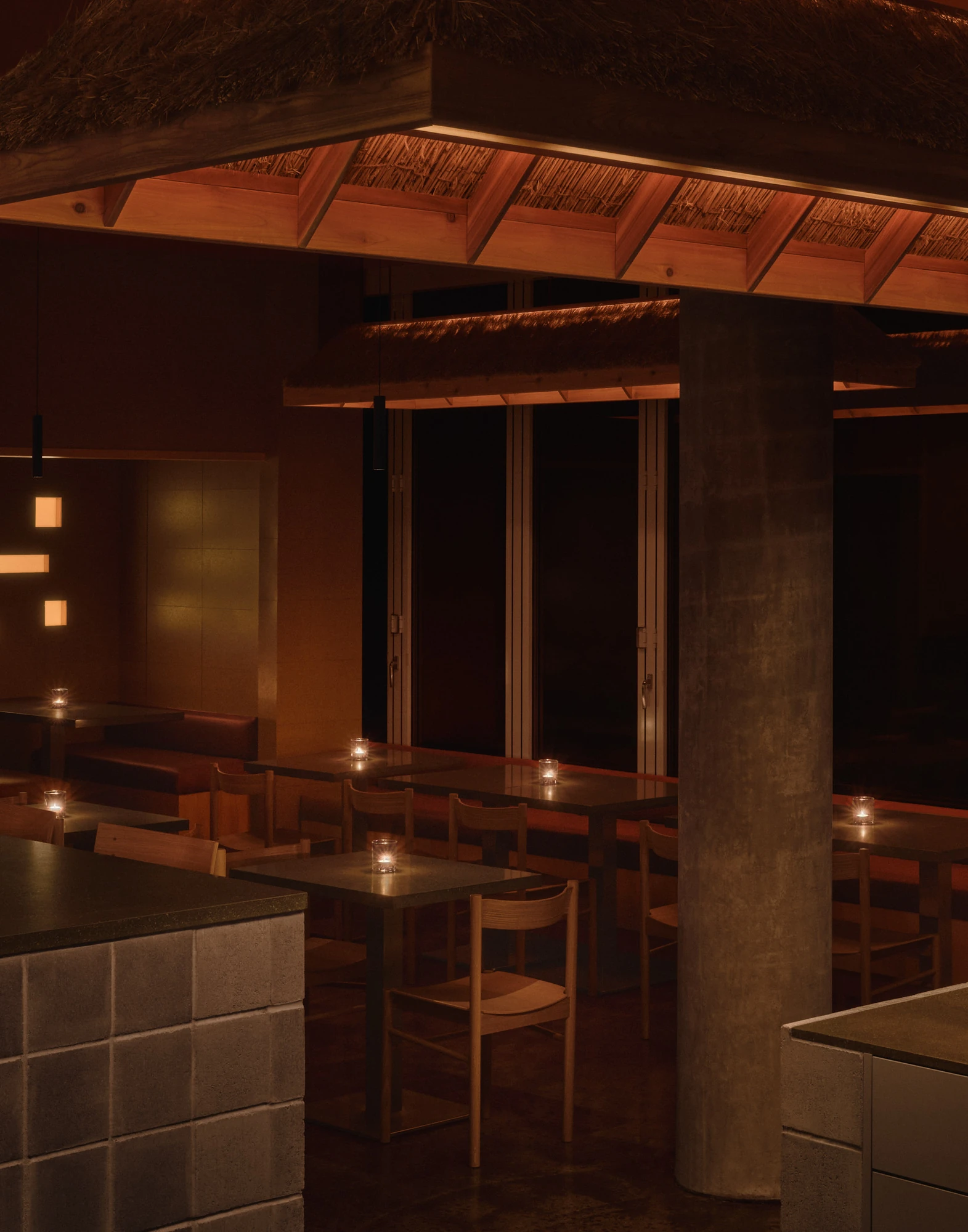 Jeju by &Daughters, photographed by Alex Lesage — Candlelit tables at night beneath a hand-thatched fixture, folding glass doors closing off the far wall.
