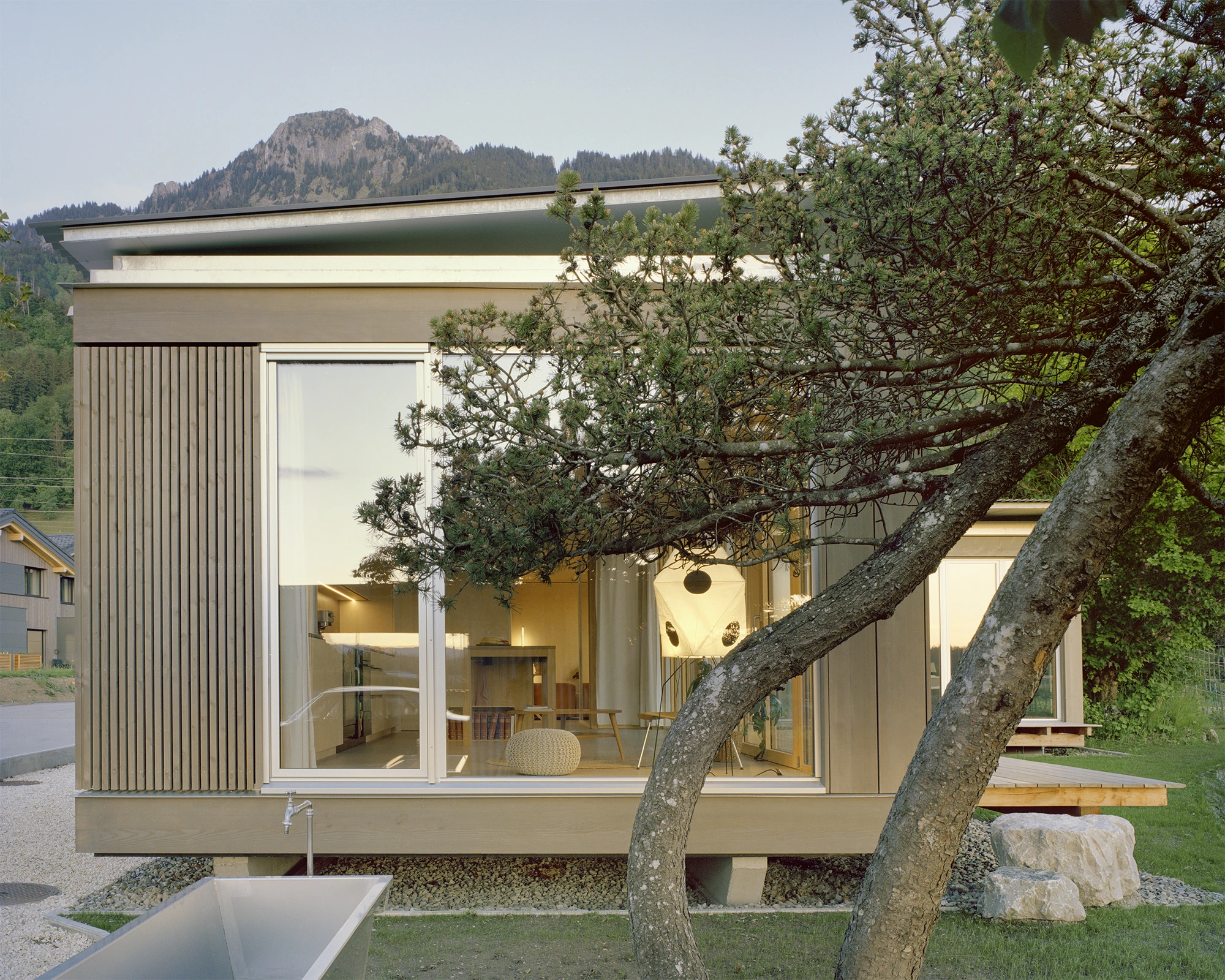 Plywood House by Charly Jolliet Architecte, photographed by Maxime Delvaux — Glazed sliding walls wrap the corner where the two timber volumes stand slightly above the preserved ground.