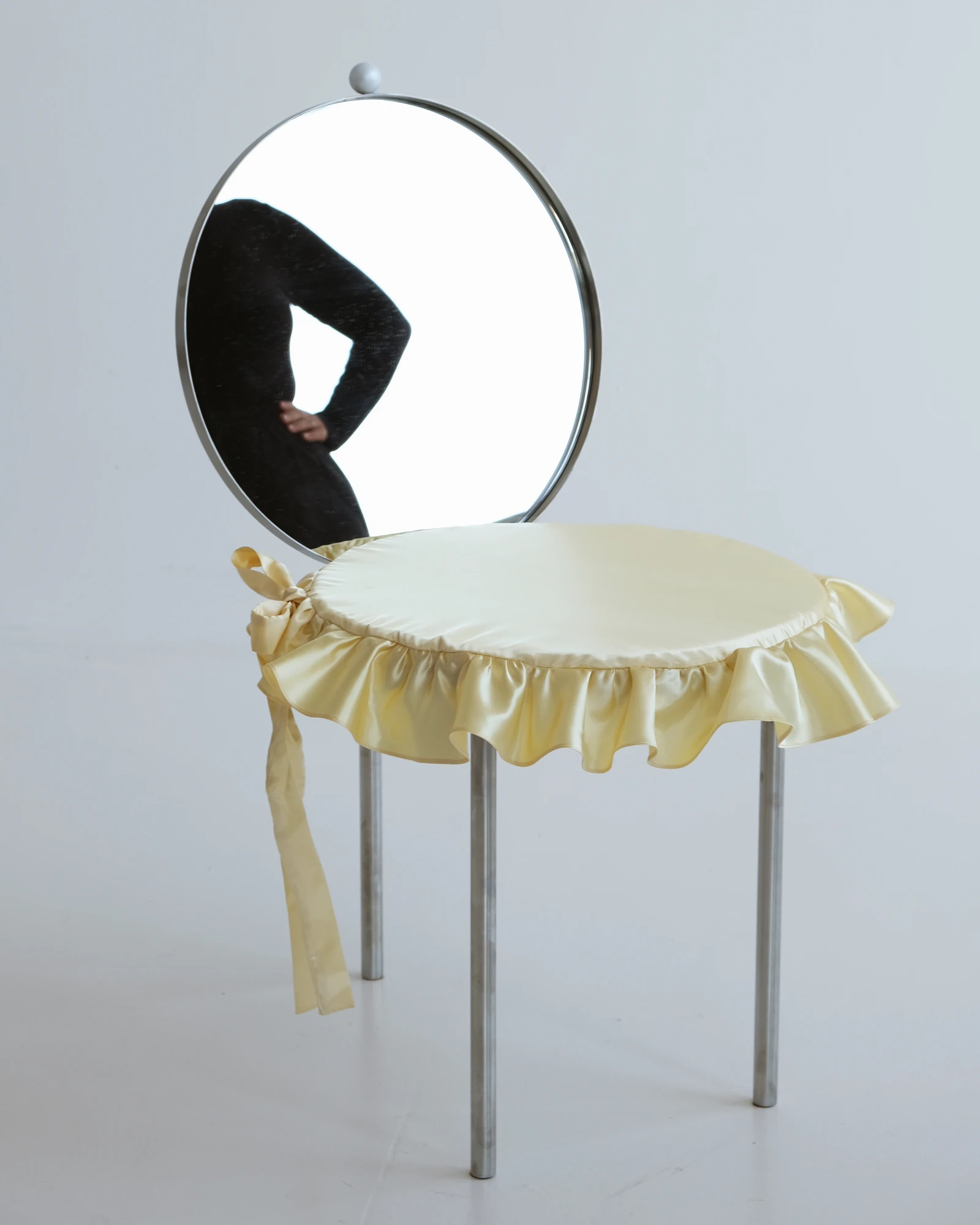 The Woman's Chair by Shushana Khachatrian, photographed by Katie Kutuzova — The round mirror backrest reflects a standing figure in black while the seat carries a frilled satin cushion on stainless-steel legs.