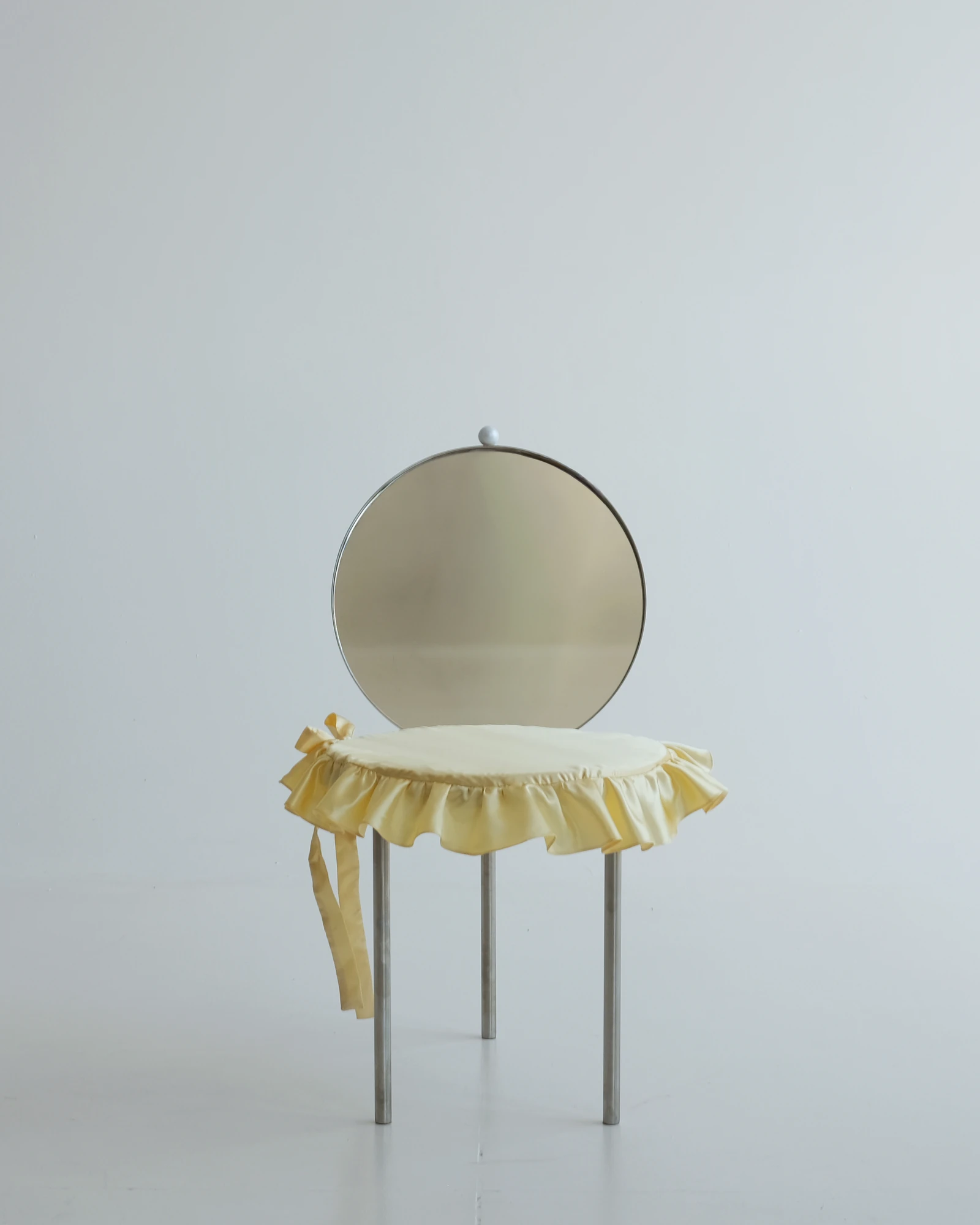 The Woman's Chair by Shushana Khachatrian, photographed by Katie Kutuzova — Front view of the chair, its seat wrapped in a frilled satin cushion tied with a ribbon below a round mirror backrest.