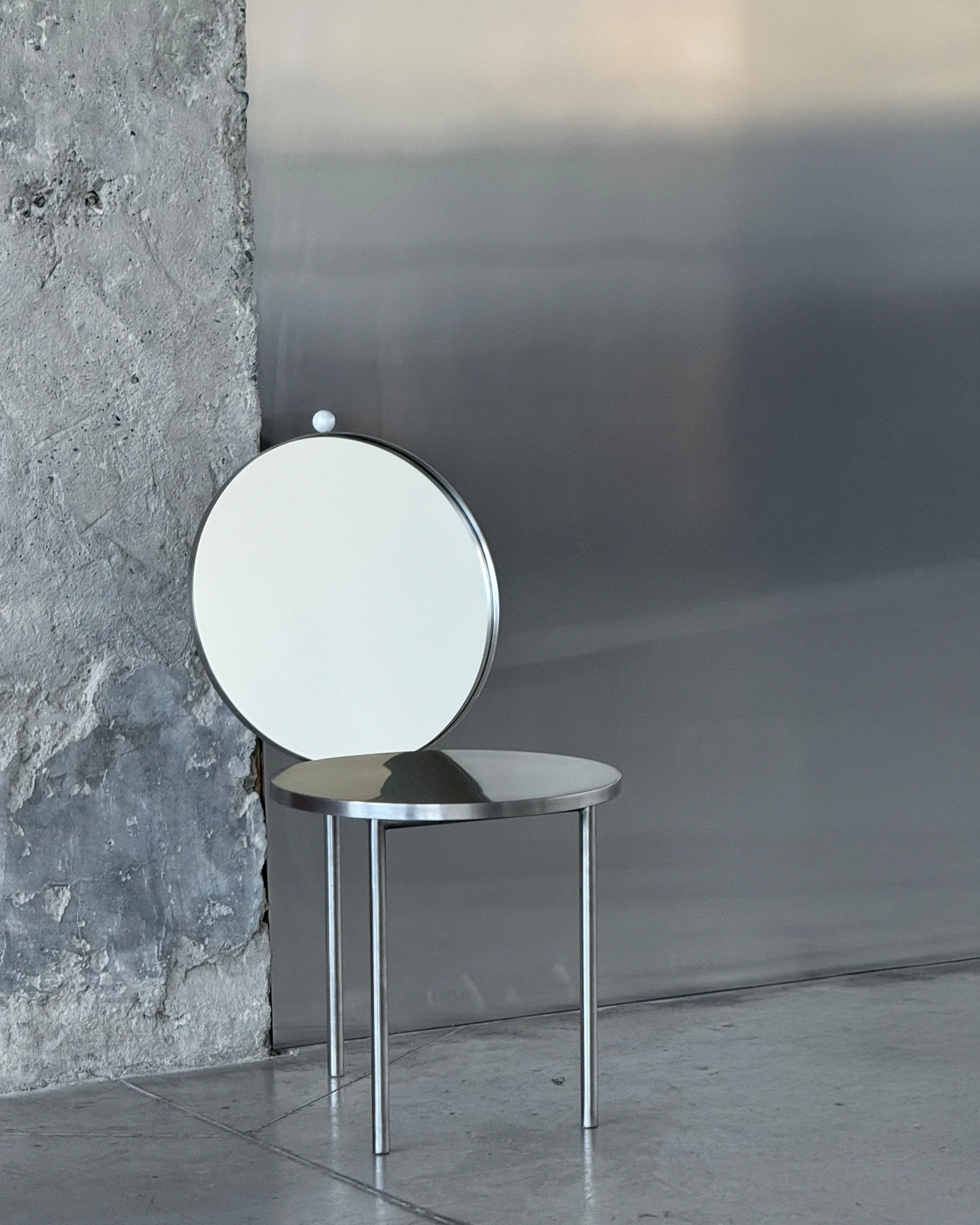 The Woman's Chair by Shushana Khachatrian, photographed by Katie Kutuzova — The bare chair against a rough concrete wall, three stainless-steel legs under a round seat and an upright mirror backrest.