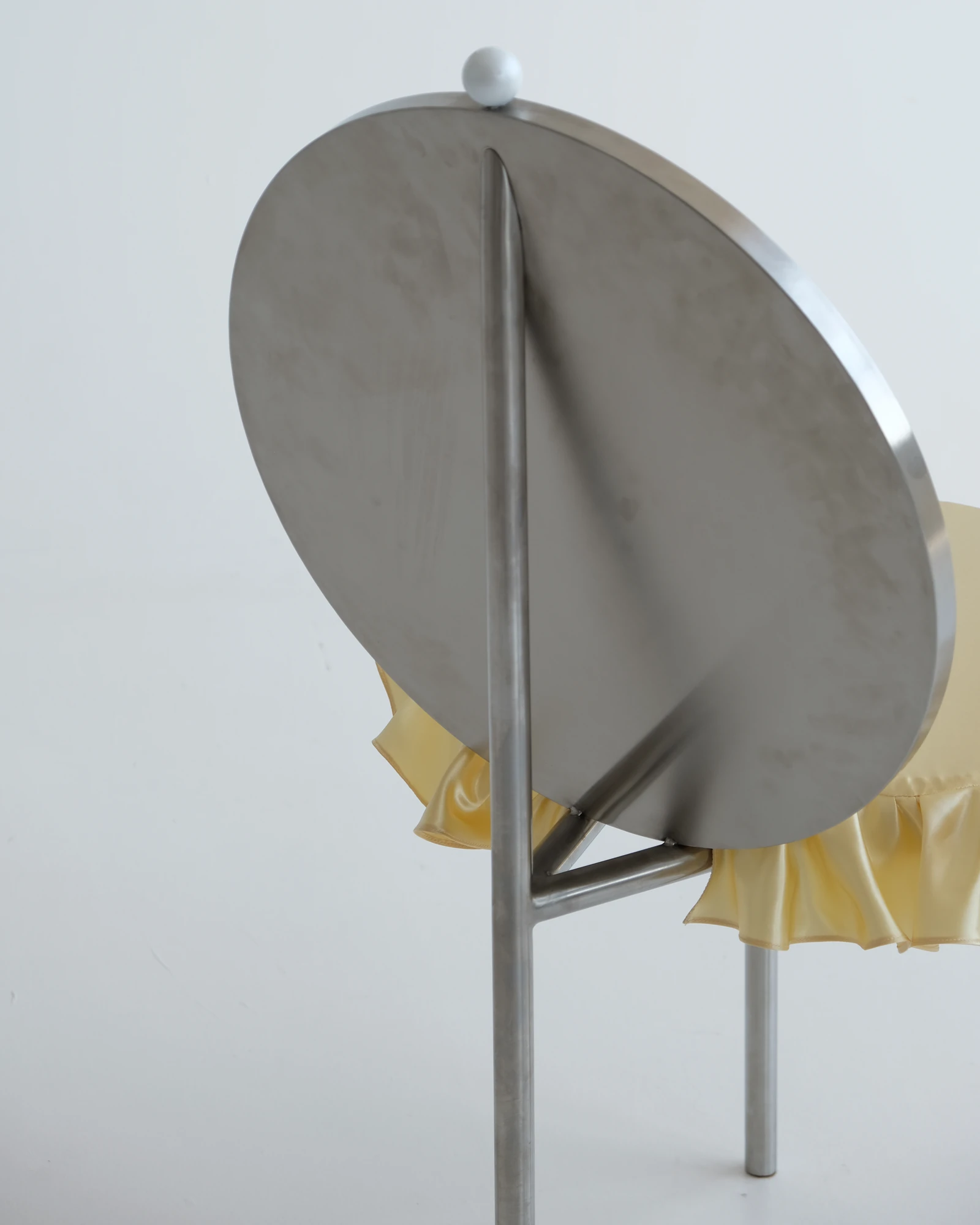 The Woman's Chair by Shushana Khachatrian, photographed by Katie Kutuzova — The back of the angled stainless-steel backrest, the frilled satin cushion visible behind and a single leg rising through the frame.