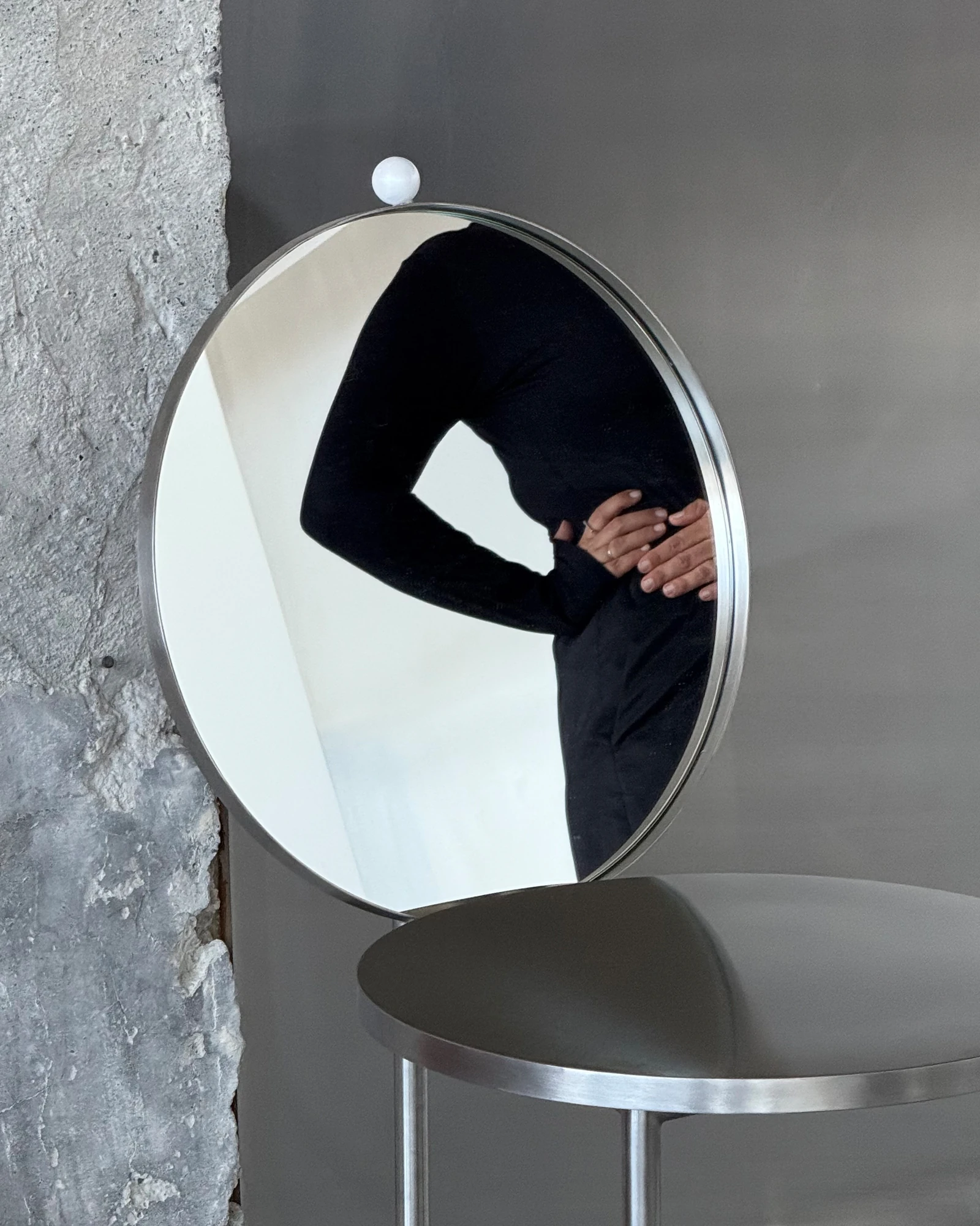 The Woman's Chair by Shushana Khachatrian, photographed by Katie Kutuzova — A round mirror backrest reflecting a figure in black, the stainless-steel seat below set against a concrete wall.