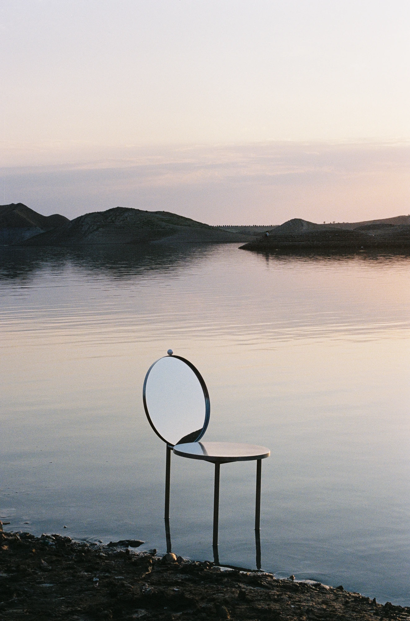 The Woman's Chair by Shushana Khachatrian, photographed by Katie Kutuzova — The chair stands on three stainless-steel legs at the edge of still water, its round mirror backrest catching the low light.