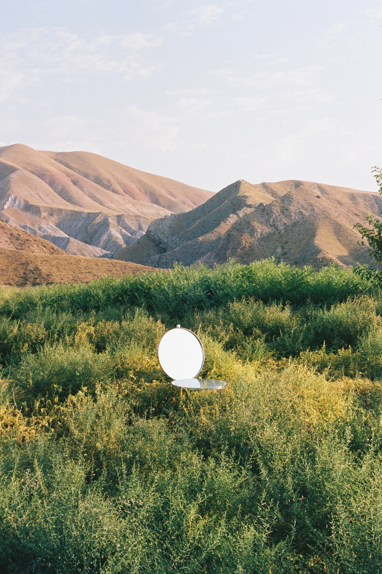 The Woman's Chair by Shushana Khachatrian, photographed by Katie Kutuzova — The chair standing in green scrub below bare mountains, its round mirror backrest bright against the foliage.