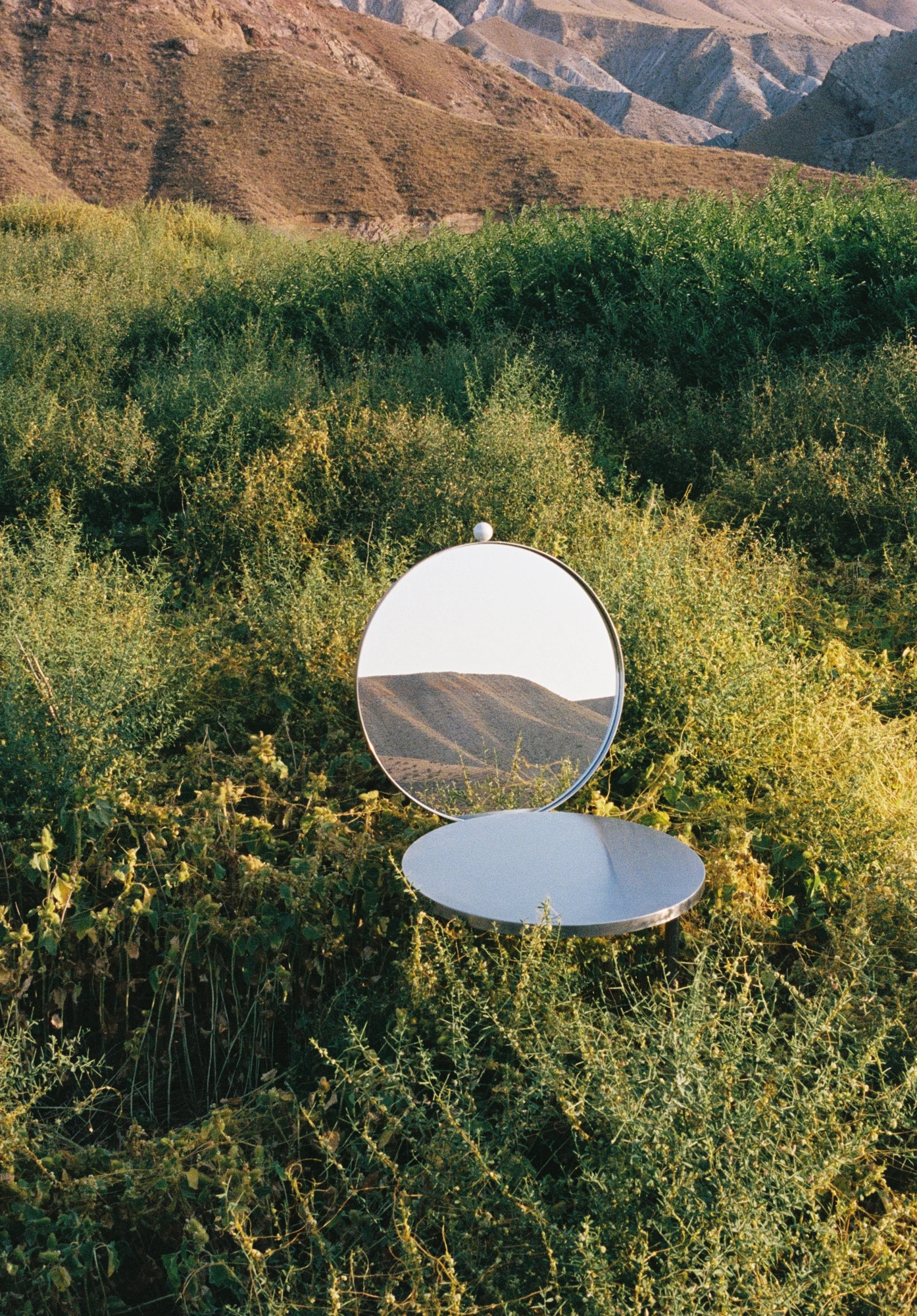 The Woman's Chair by Shushana Khachatrian, photographed by Katie Kutuzova — A close view of the chair outdoors, the round mirror backrest and reflective steel seat set among wild green growth.