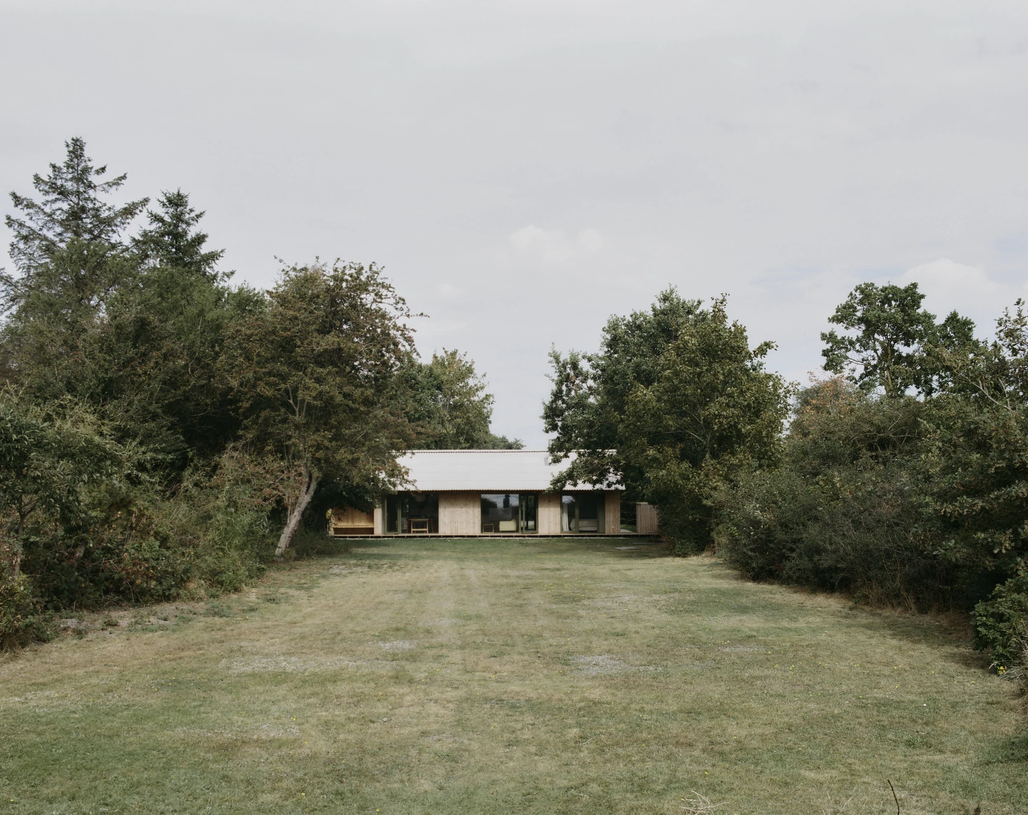 Stenkløvervej Vacation House by Studio Furö, photographed by Ditte Auguste Mørkholt — The house at the end of a mown clearing, framed by trees on either side under an overcast sky.