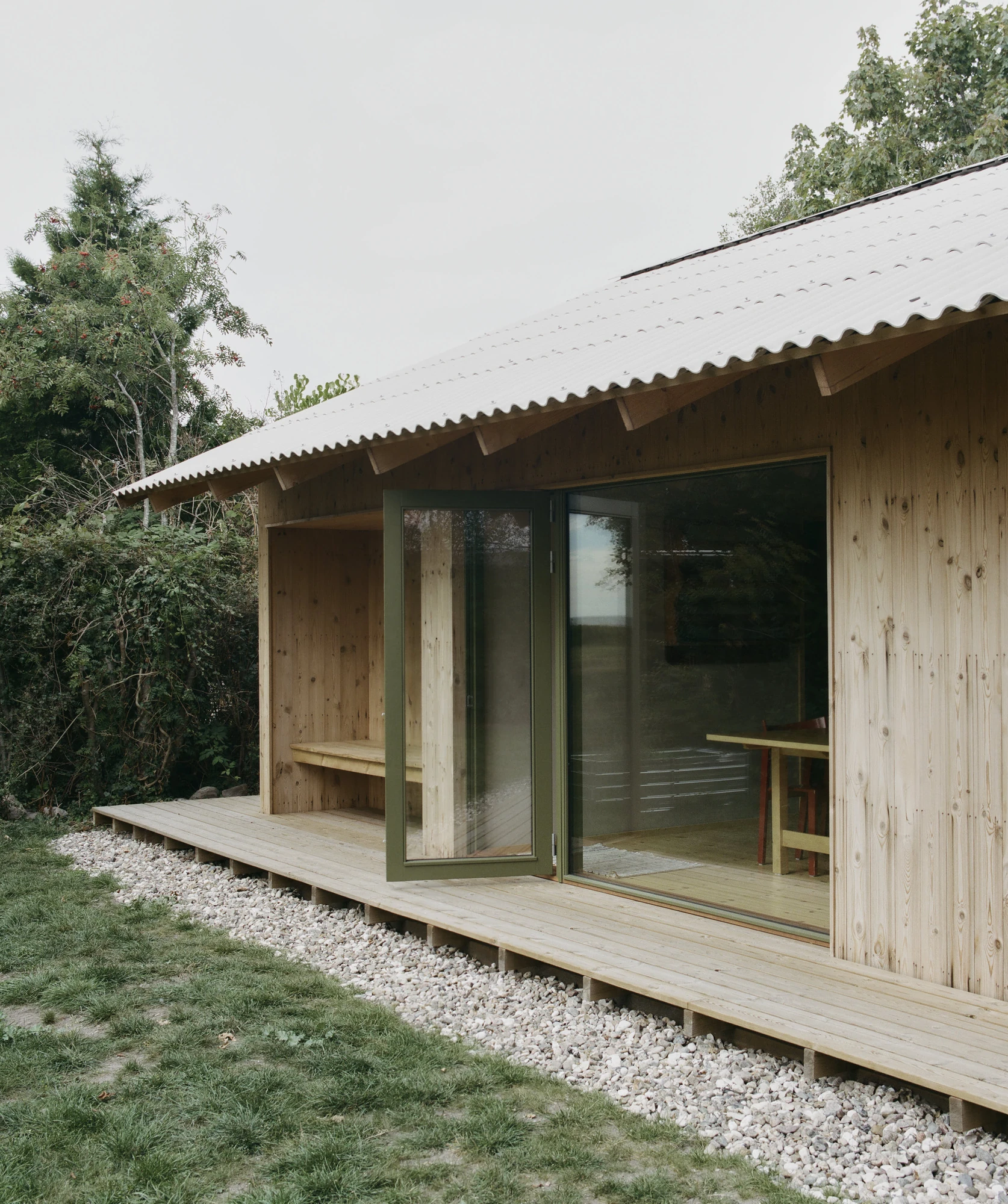 Stenkløvervej Vacation House by Studio Furö, photographed by Ditte Auguste Mørkholt — A corner of the house where the roof overhangs a recess in the deck, an open door beside it.