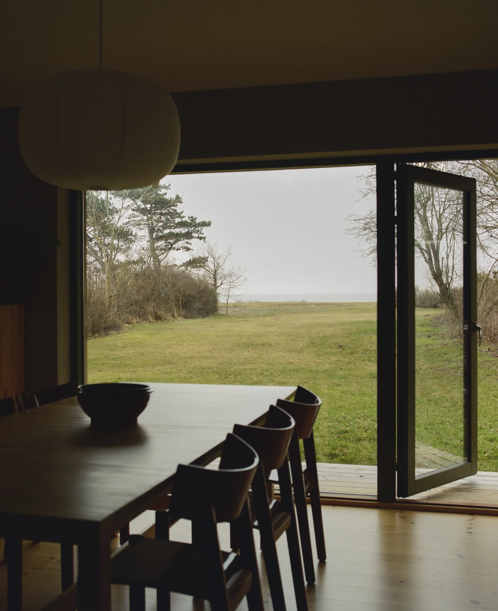 Stenkløvervej Vacation House by Studio Furö, photographed by Ditte Auguste Mørkholt — The view from the dining table through open doors to a field and the water beyond it.