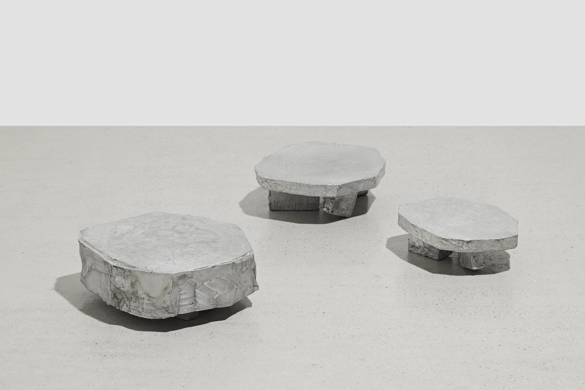 Lying on the floor by StudioFA, photographed by Yongin Myoung — Three low cast aluminum forms stand apart on a pale floor against a white wall, each a faceted slab raised on short legs.