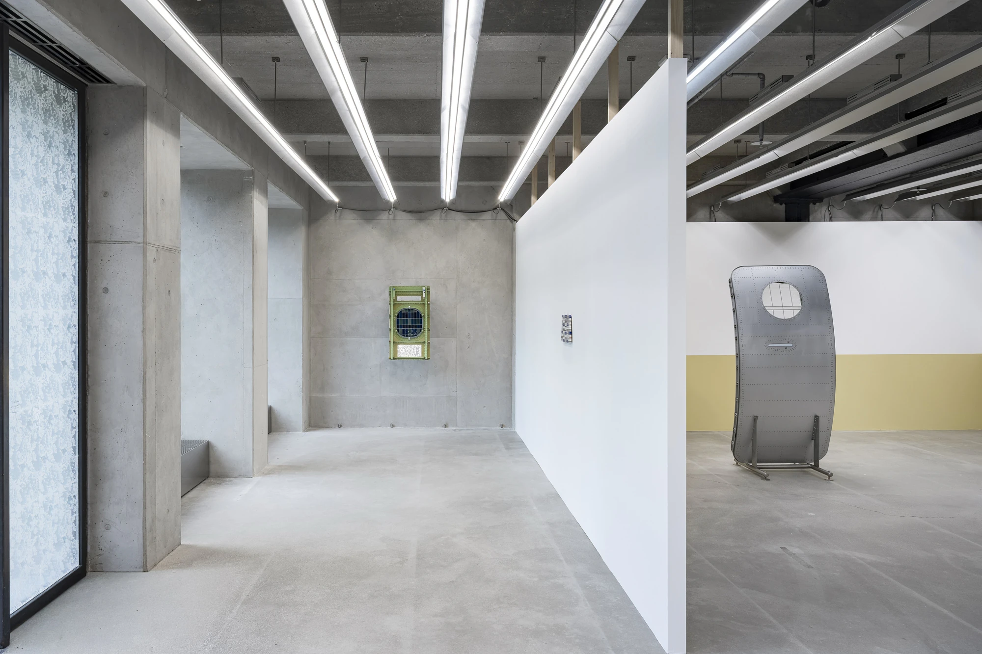 Addresses Without Names by Ju Young Kim, photographed by Dirk Tacke — Installation view of addresses without names at max goelitz, Munich, a lace-printed glass panel at the window, a green-framed aircraft window on the concrete wall and a riveted…