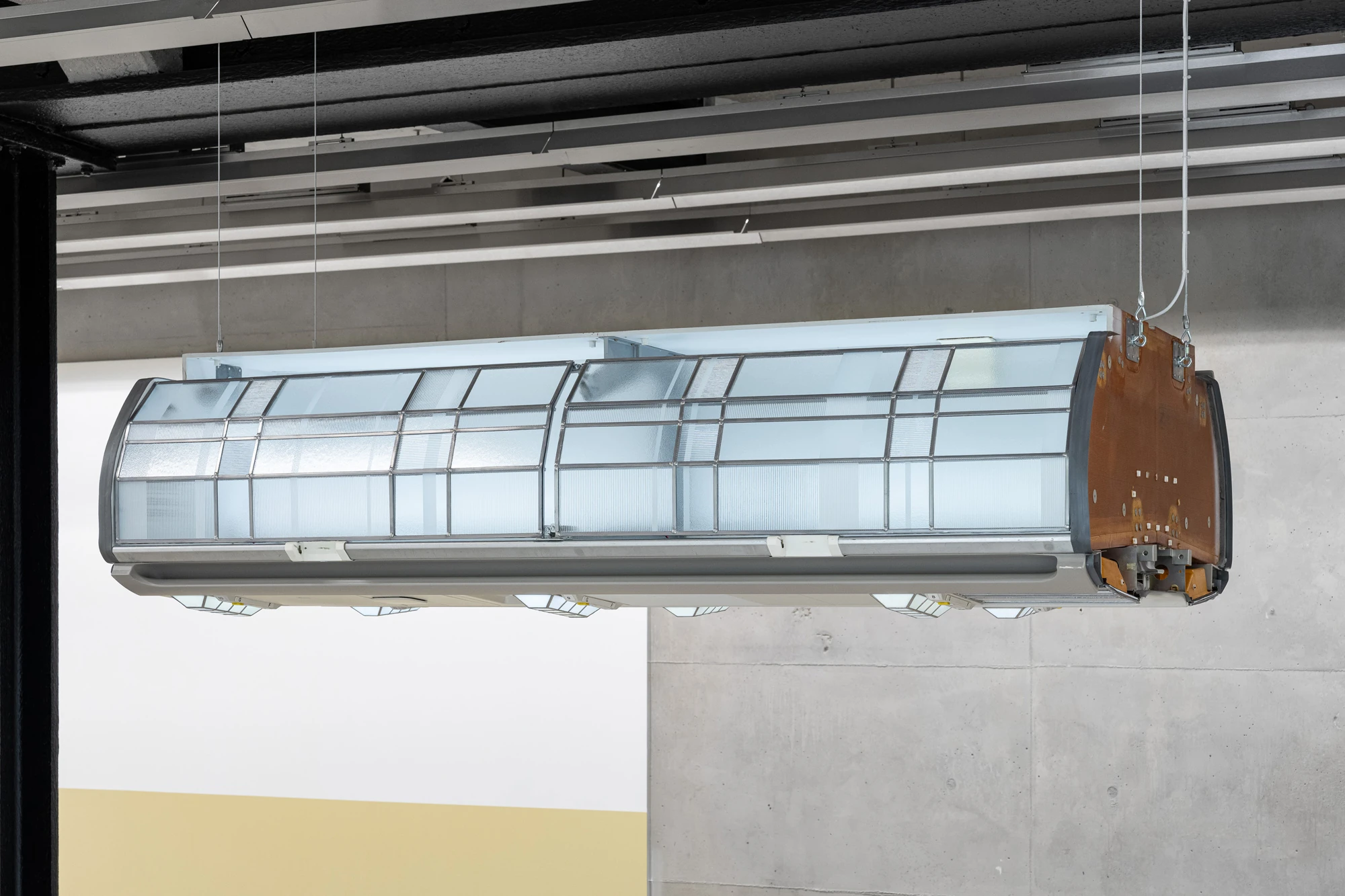 Addresses Without Names by Ju Young Kim, photographed by Dirk Tacke — She slept better when nothing stayed still, 2026, an aircraft overhead compartment rebuilt in leaded and frosted glass with an amber-brown end and lit from within, 55 x 212 x 130 cm.
