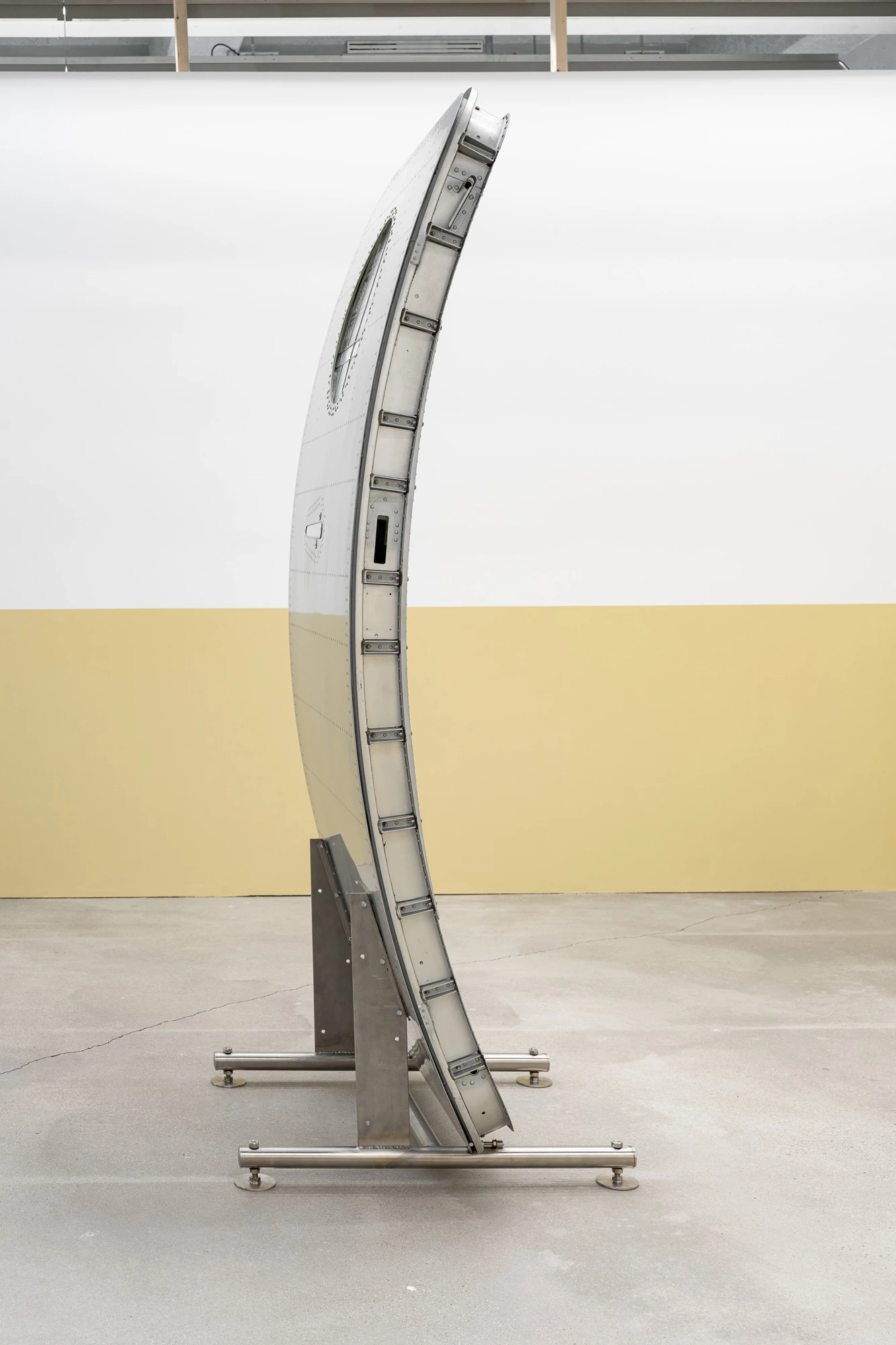 Addresses Without Names by Ju Young Kim, photographed by Dirk Tacke — Names behind the sun-faded plastic, 2026, a curved aircraft exterior door standing on a steel base with half its skin painted ochre, 200 x 92 x 78 cm.