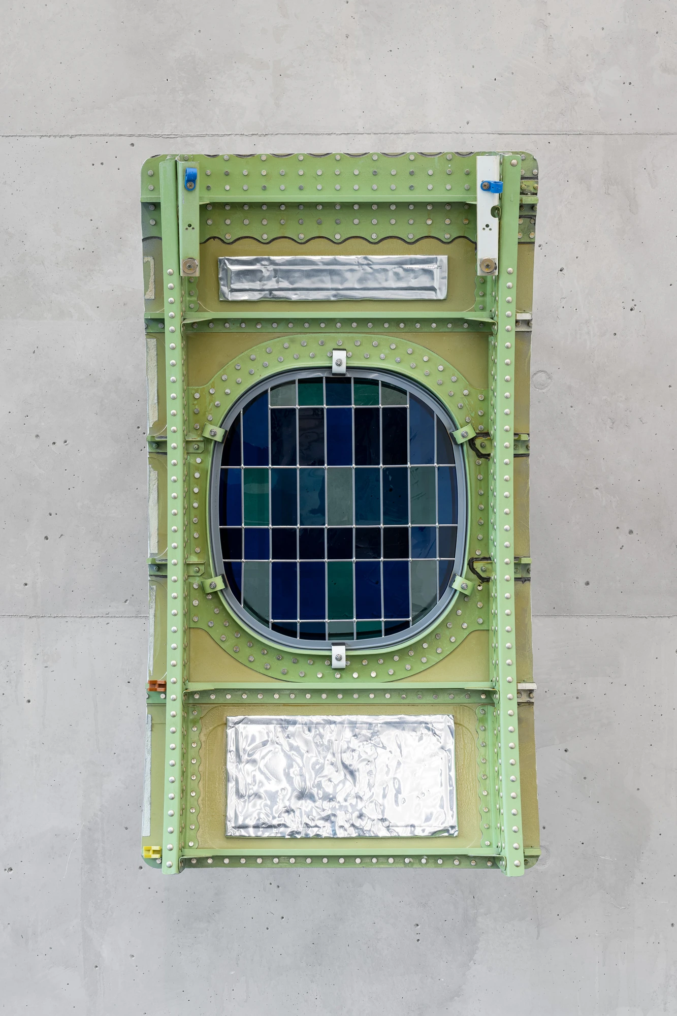 Addresses Without Names by Ju Young Kim, photographed by Dirk Tacke — She always sat on the same side of the table, 2026, an aircraft exterior window in green riveted metal holding an oval of colored and sanded glass assembled with lead and tin on…