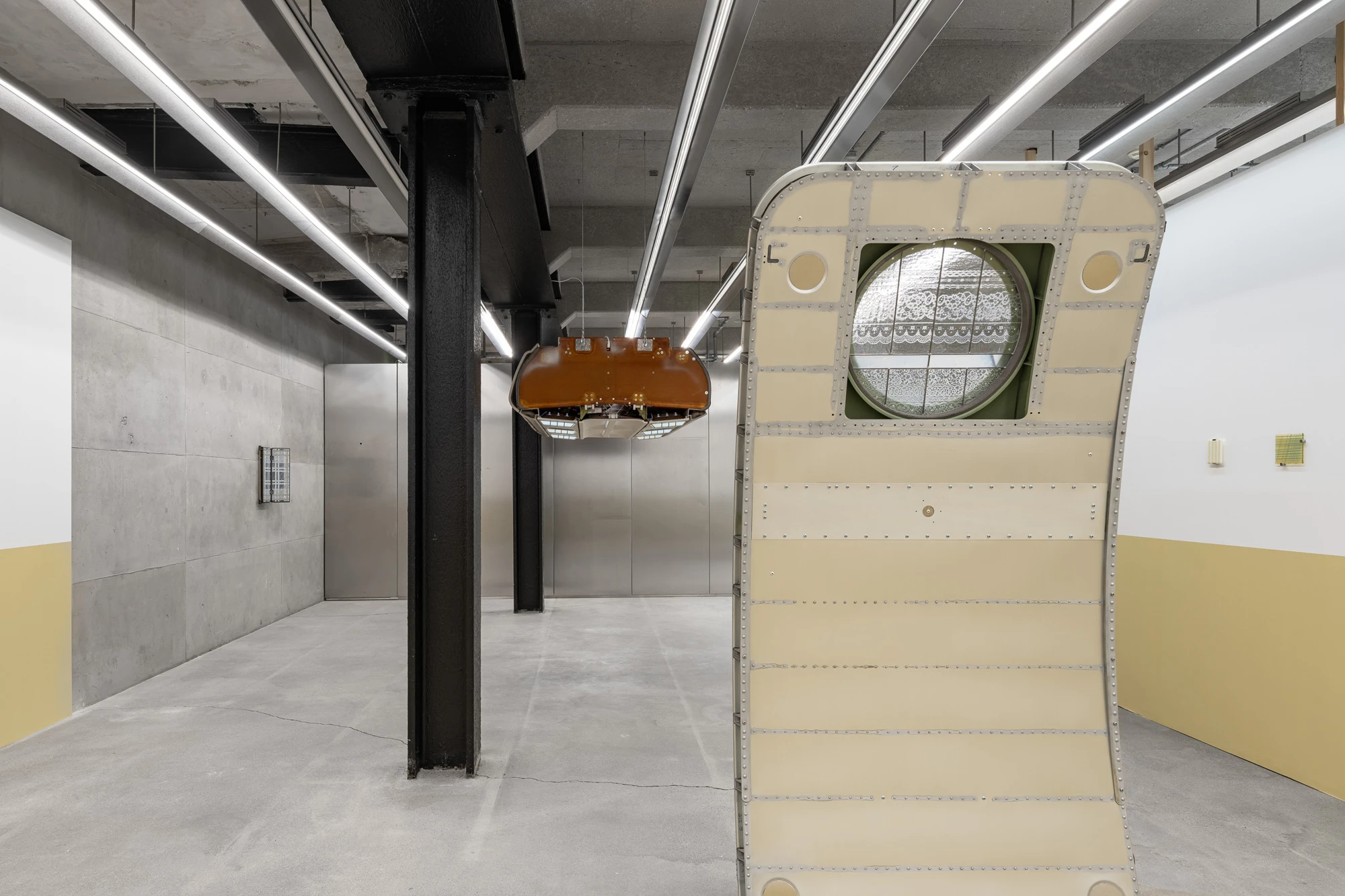 Addresses Without Names by Ju Young Kim, photographed by Dirk Tacke — Installation view with the cream riveted aircraft door and its lit lace-printed window in the foreground and the overhead compartment hanging in the concrete hall beyond.