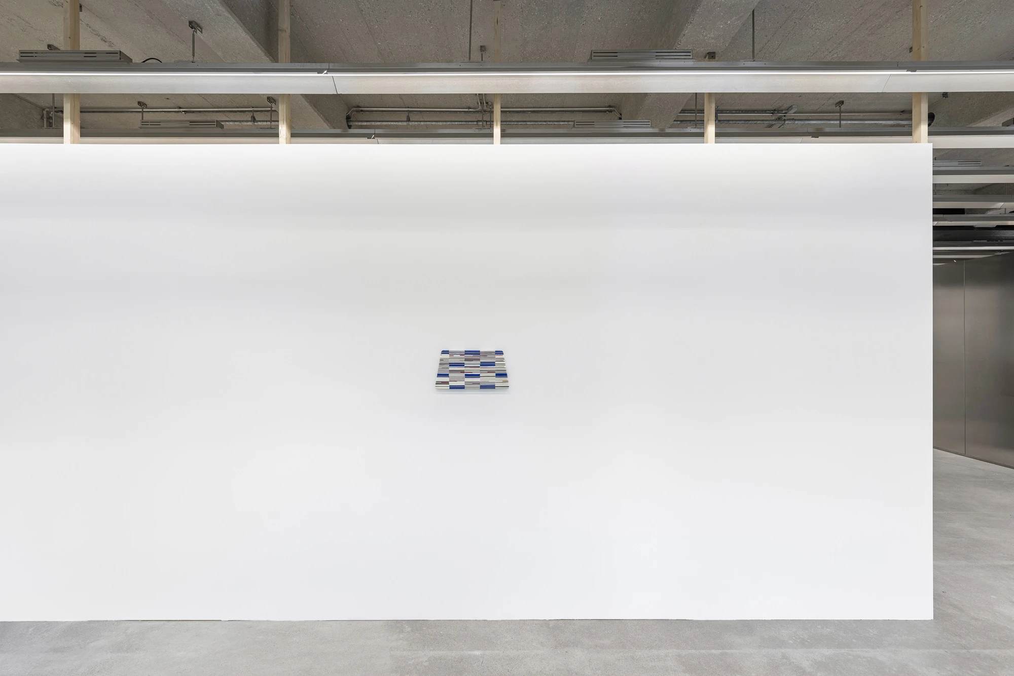 Addresses Without Names by Ju Young Kim, photographed by Dirk Tacke — Installation view of a small blue, grey and white checkerboard glass panel centered on a large white wall.