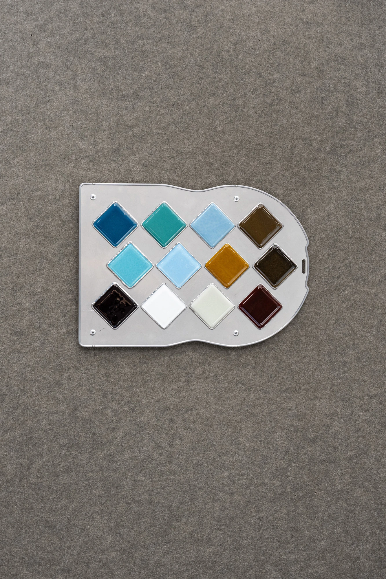 Addresses Without Names by Ju Young Kim, photographed by Dirk Tacke — She always left the blue unused, 2026, twelve squares of colored glass in blues, amber and brown set into a clear fused-glass panel like a boxed paint set, assembled with lead and tin…