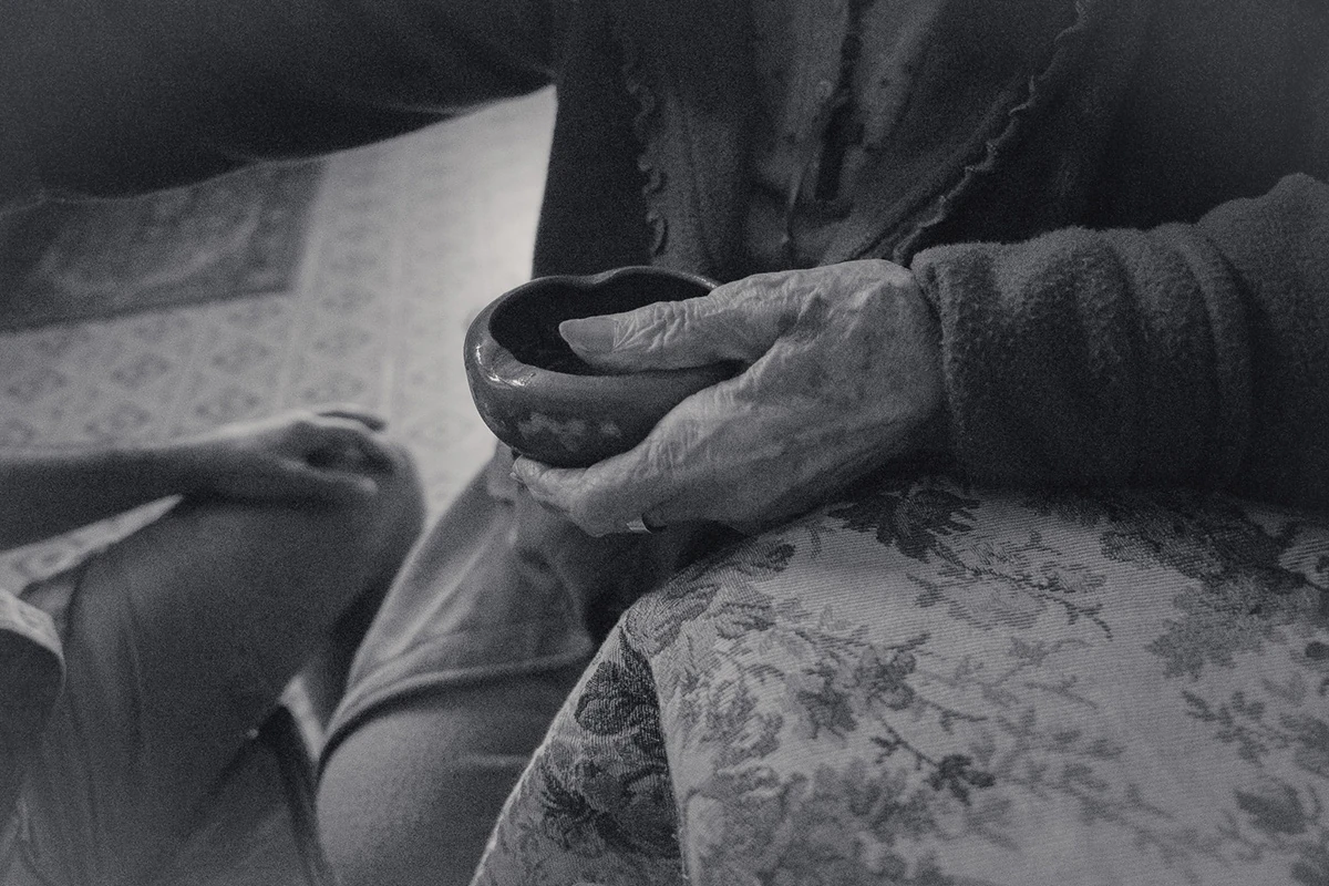 An Island Within the Island by succco, photographed by succco — Weathered hands cradle a small dark ceramic bowl in the lap of a seated figure, resting on floral upholstery above a patterned tile floor.