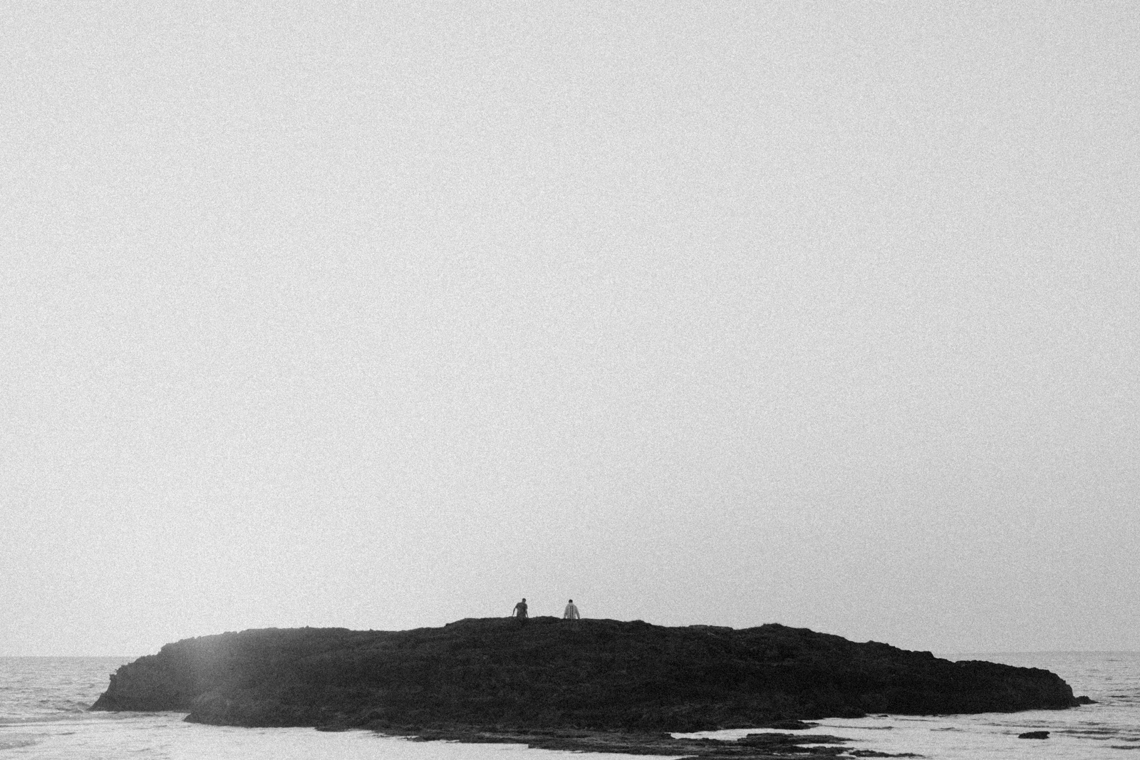 An Island Within the Island by succco, photographed by succco — Two small figures stand on the dark ridge of a low rocky islet in a hazy sea, the pale sky filling most of the frame.