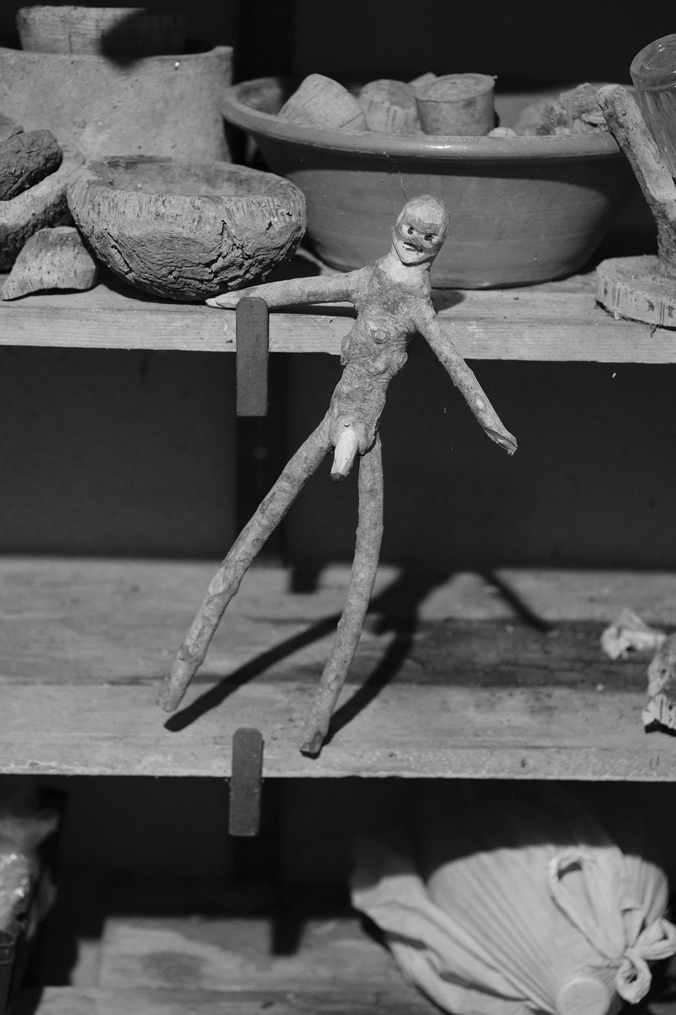 An Island Within the Island by succco, photographed by succco — A crudely carved figure of root and stick is propped on a wooden shelf among earthen bowls and vessels.
