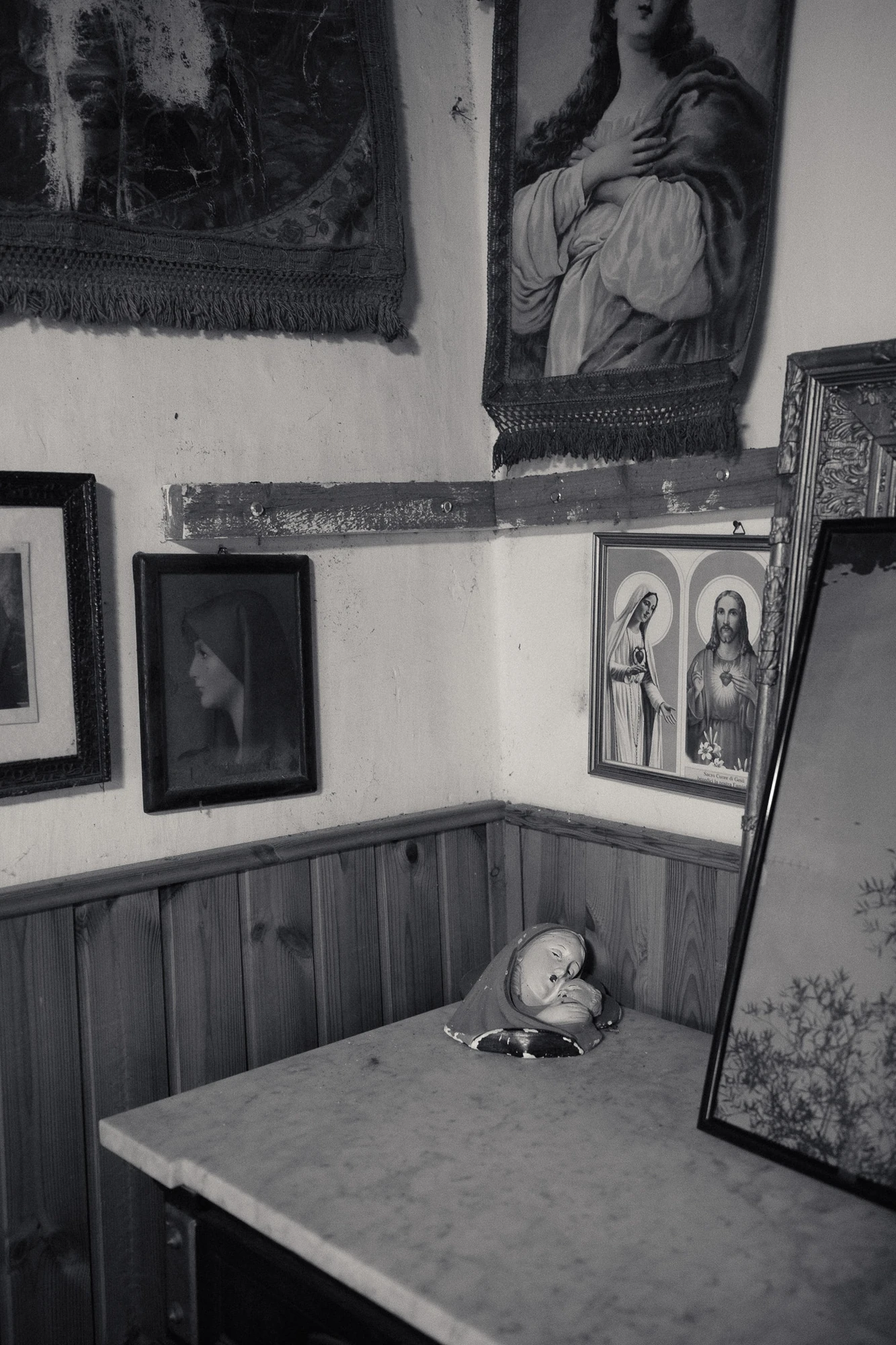 An Island Within the Island by succco, photographed by succco — A domestic corner crowded with framed devotional prints and embroidered banners, a small plaster Madonna-and-child figurine laid on a marble-topped chest.