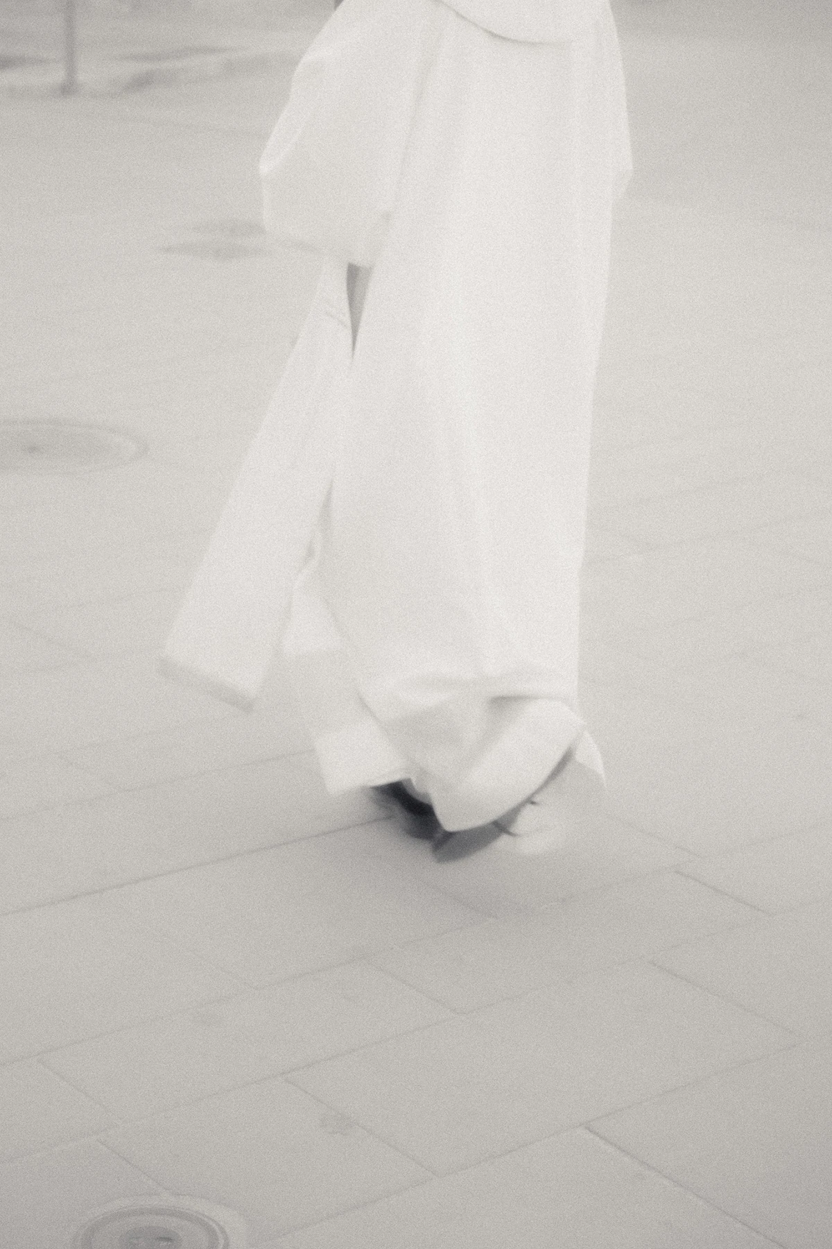 An Island Within the Island by succco, photographed by succco — A figure in a long, loose white garment walks across a pale tiled floor, cropped below the waist, the frame nearly overexposed.