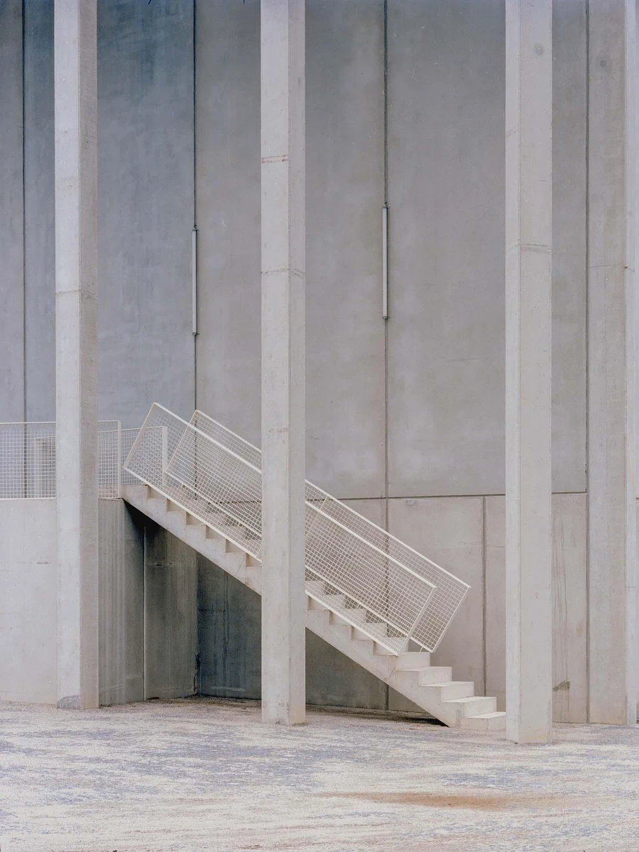 Corcet Headquarters by Nuno M. Sousa, photographed by Federico Farinatti, Francisco Ascensão — An external stair with a mesh balustrade cuts diagonally behind a slender concrete pillar, framed tightly against a plain concrete wall.