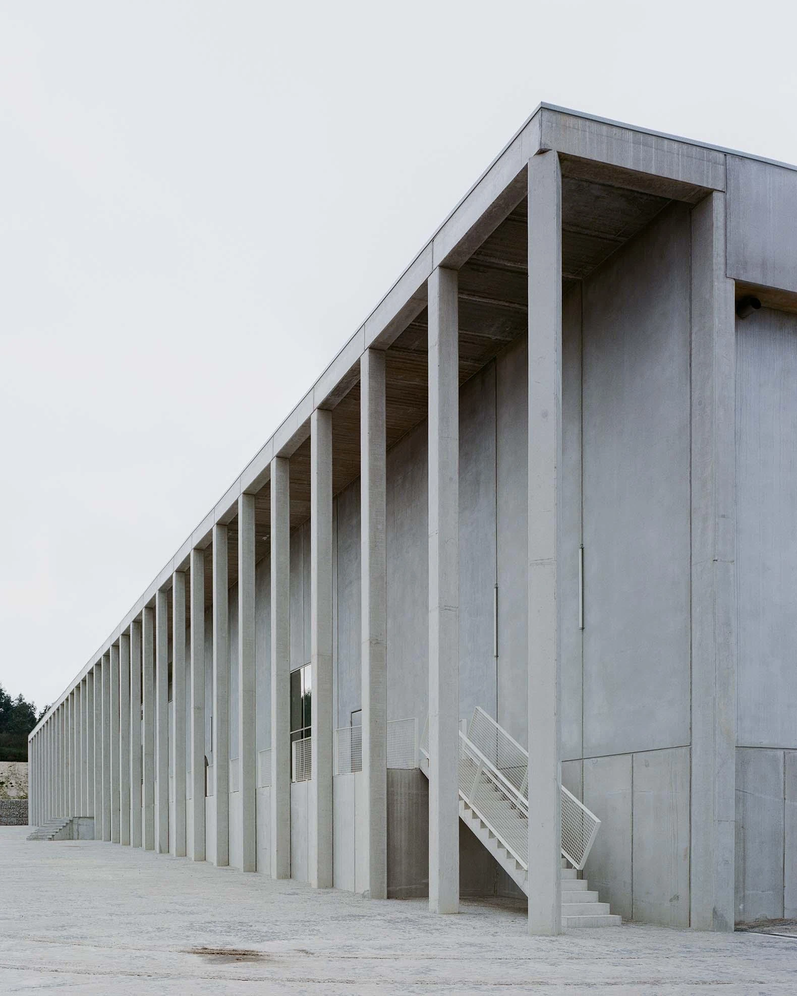 Corcet Headquarters by Nuno M. Sousa, photographed by Federico Farinatti, Francisco Ascensão — The building turns a corner under a continuous run of tall concrete pillars, an external stair dropping to the paved ground at the near end.