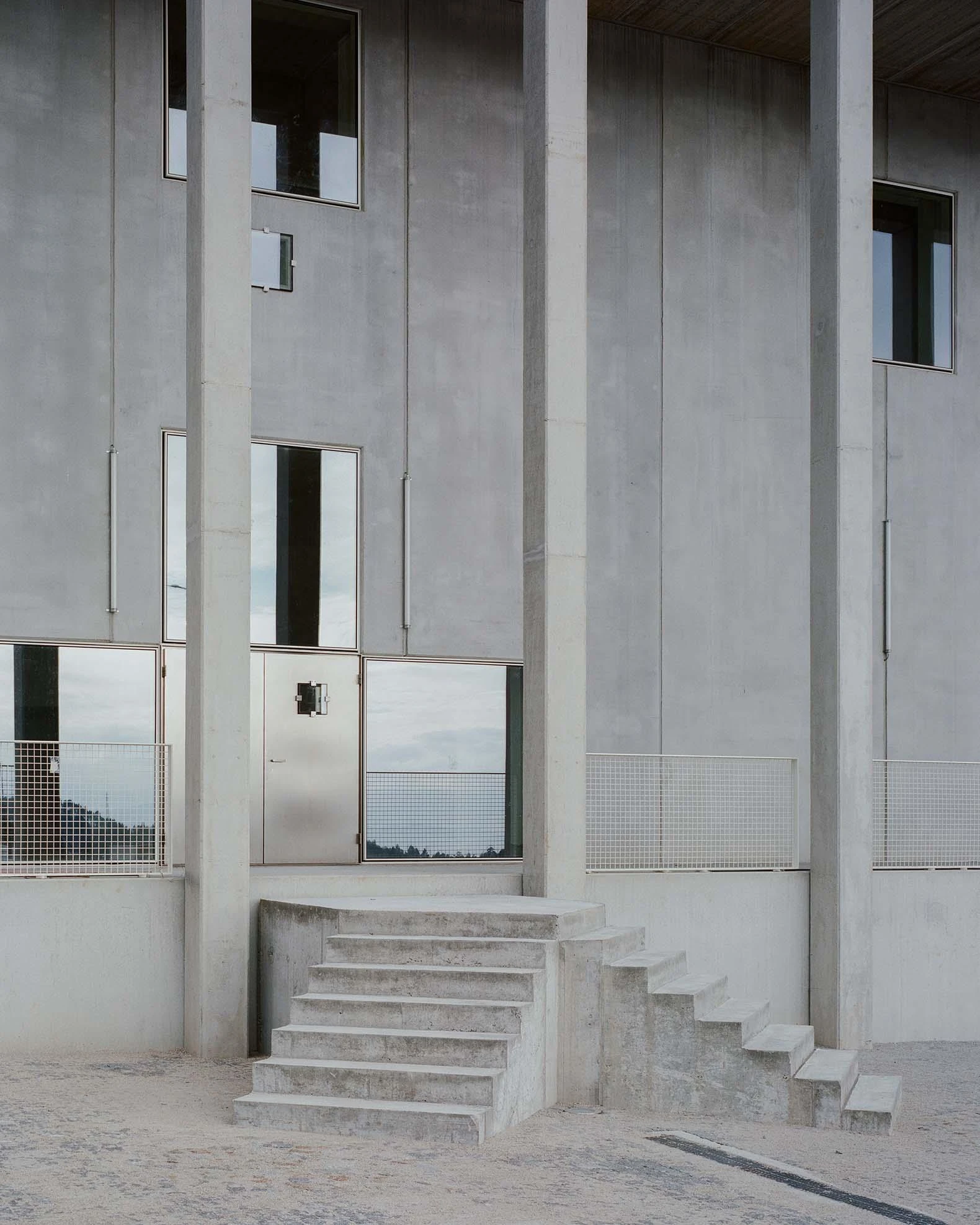 Corcet Headquarters by Nuno M. Sousa, photographed by Federico Farinatti, Francisco Ascensão — Wide concrete steps climb between two tall pillars to a glazed entrance, the panes mirroring a pale sky above the raised threshold.