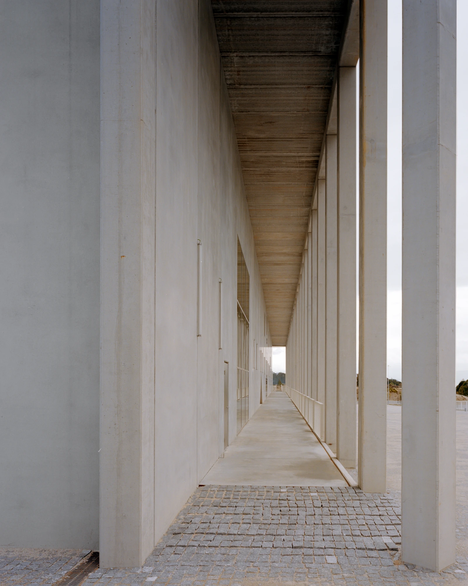 Corcet Headquarters by Nuno M. Sousa, photographed by Federico Farinatti, Francisco Ascensão — The covered gallery runs to a bright vanishing point, cobbled paving to one side and concrete underfoot, daylight slipping between the pillars.