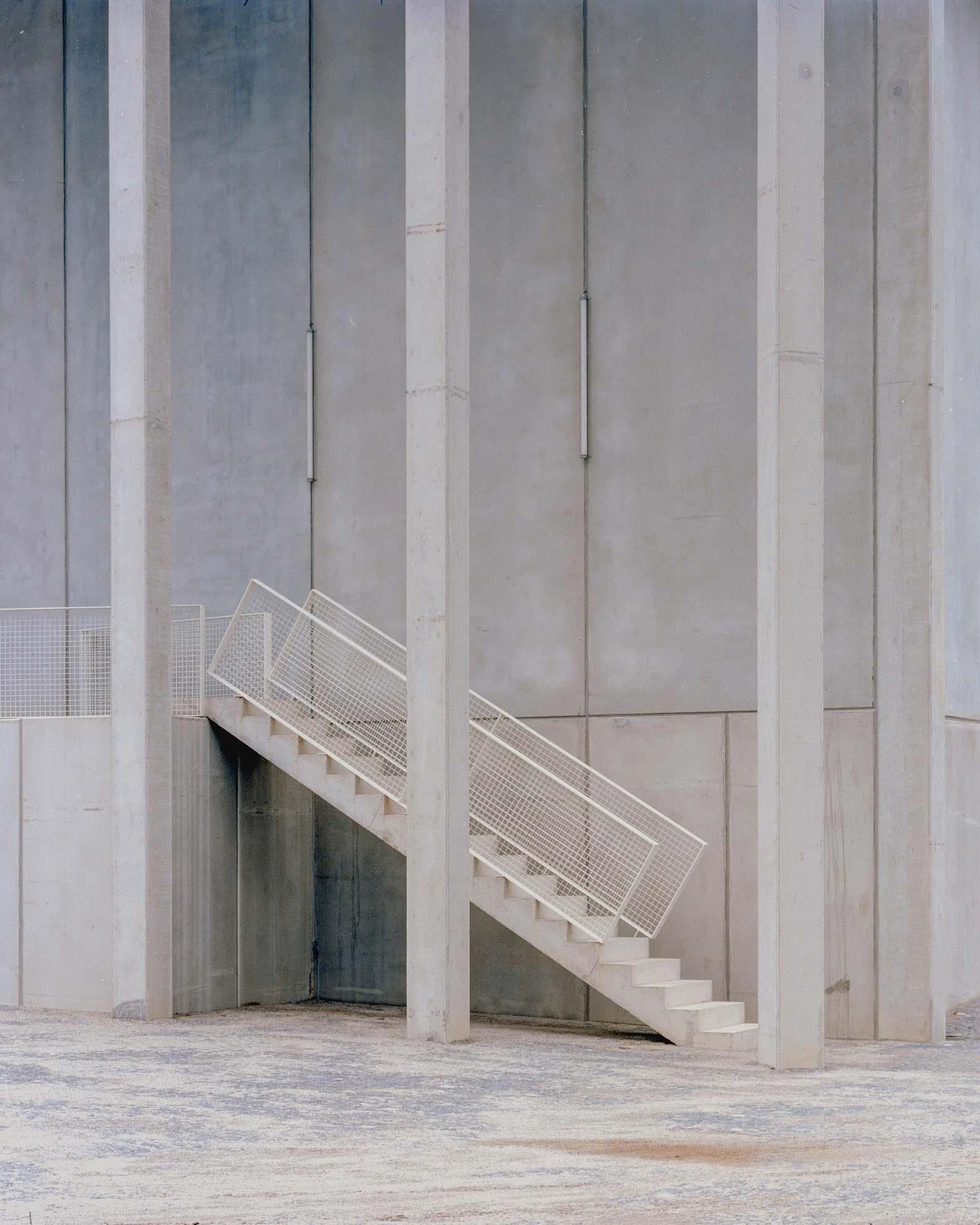 Corcet Headquarters by Nuno M. Sousa, photographed by Federico Farinatti, Francisco Ascensão — A slender concrete pillar crosses in front of an external stair, its mesh balustrade catching the light against a plain concrete wall.