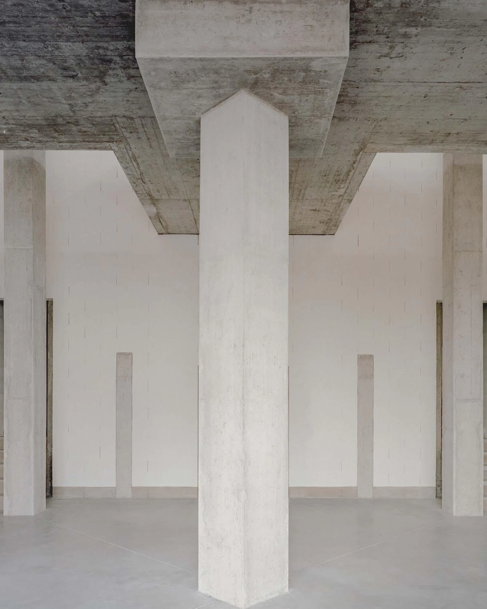 Corcet Headquarters by Nuno M. Sousa, photographed by Federico Farinatti, Francisco Ascensão — Looking up, a pale column widens where it meets a rough concrete soffit, flanked by tall white walls scored with narrow vertical joints.