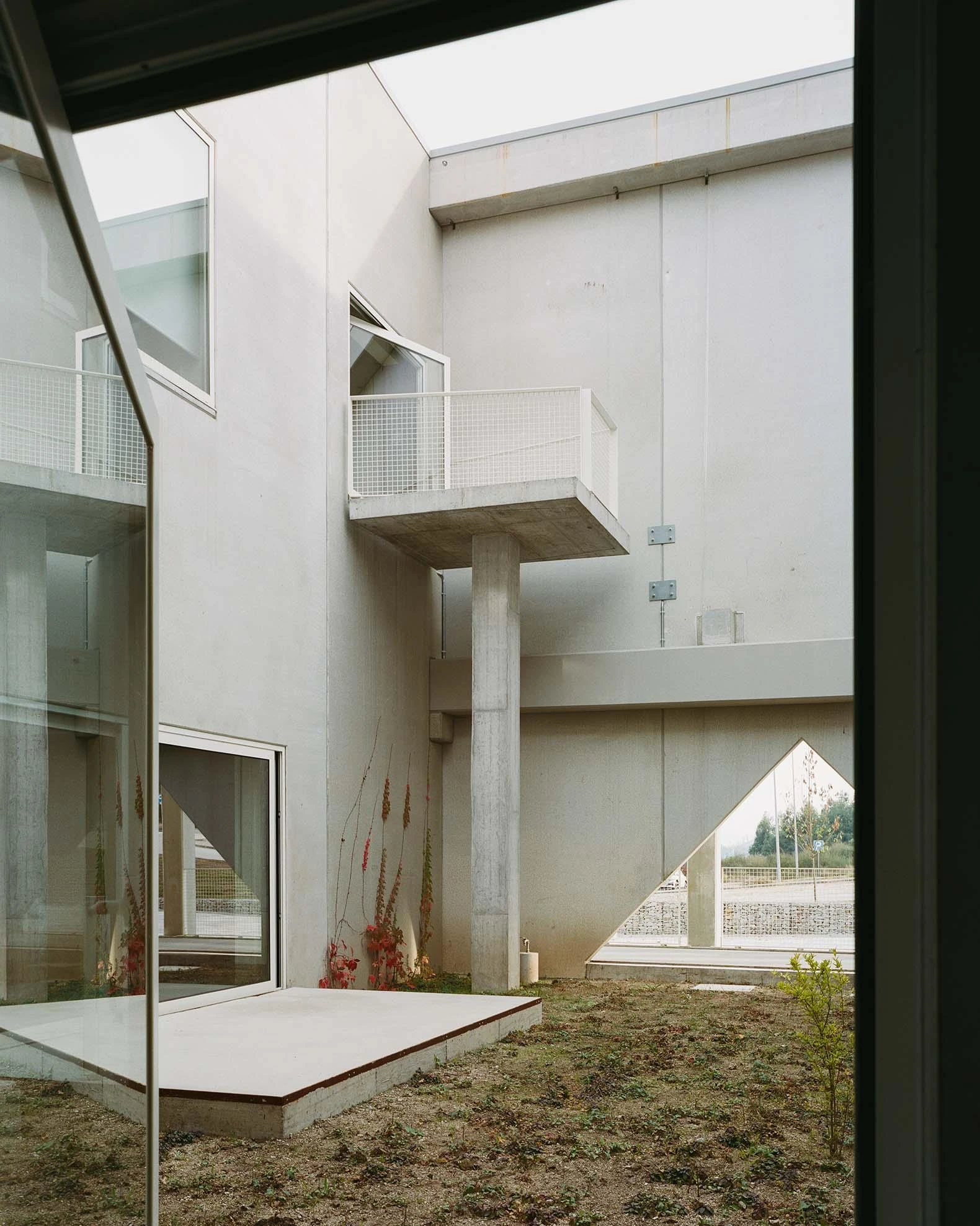 Corcet Headquarters by Nuno M. Sousa, photographed by Federico Farinatti, Francisco Ascensão — Seen through glass, the courtyard opens two storeys high, its cantilevered platform and slim pillar set against a triangular view out to the road.