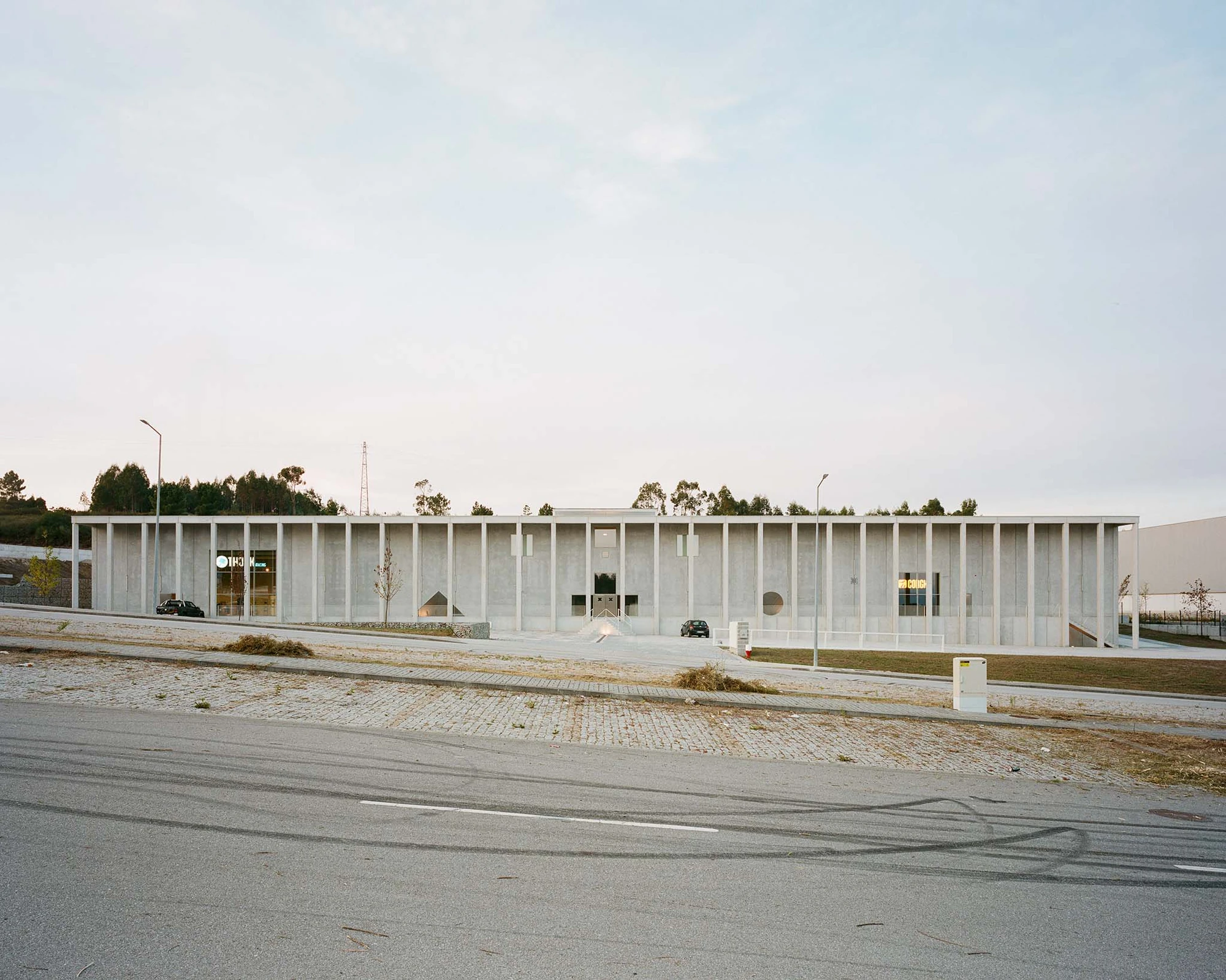 Corcet Headquarters by Nuno M. Sousa, photographed by Federico Farinatti, Francisco Ascensão — Seen from across the road, the low building presents a full-length screen of thin concrete pillars, cars parked in its shadow and a wooded hill rising behind.