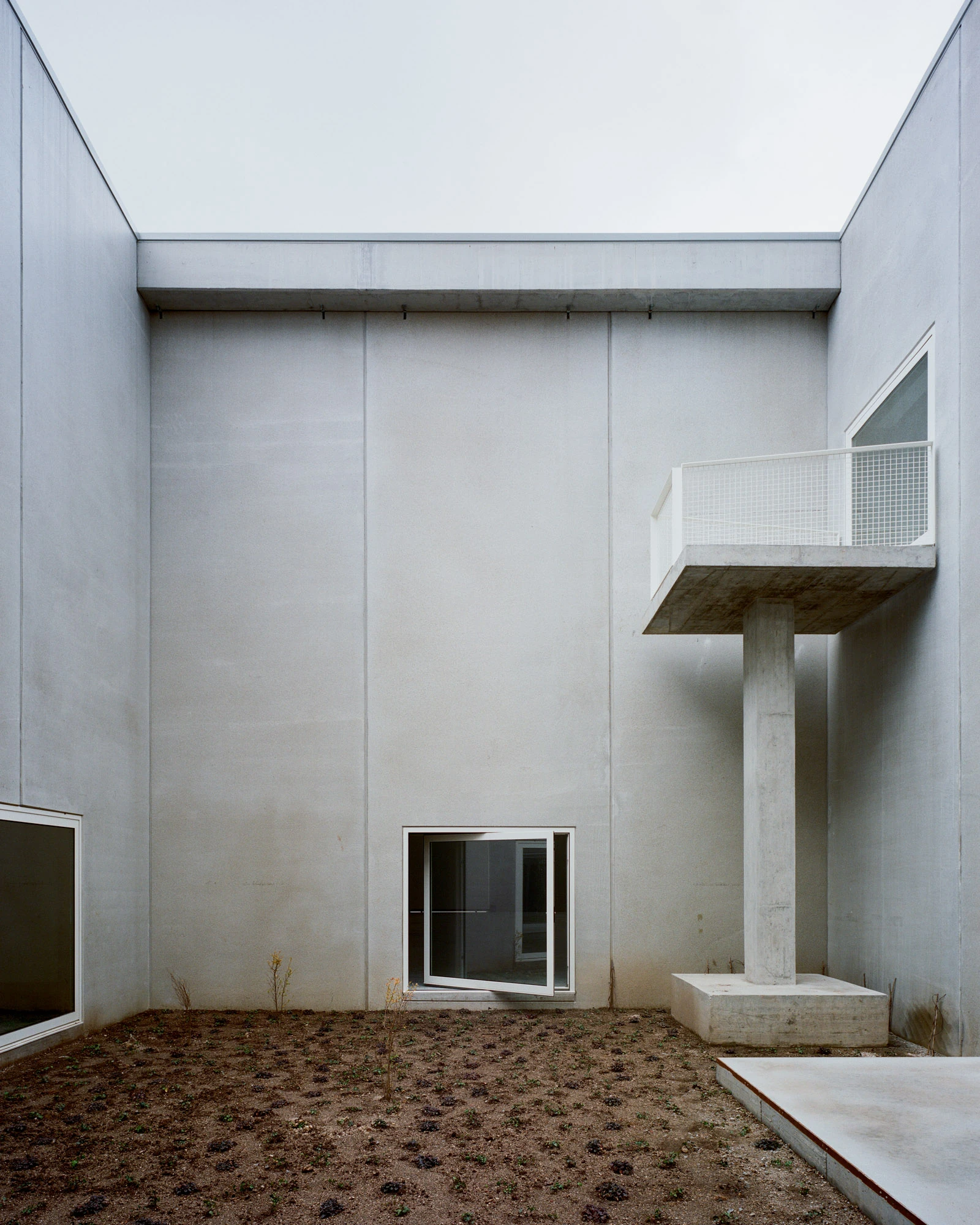 Corcet Headquarters by Nuno M. Sousa, photographed by Federico Farinatti, Francisco Ascensão — Inside a tall courtyard, a concrete balcony cantilevers from a lone pillar, a red climbing plant tracing the wall beside a triangular window.