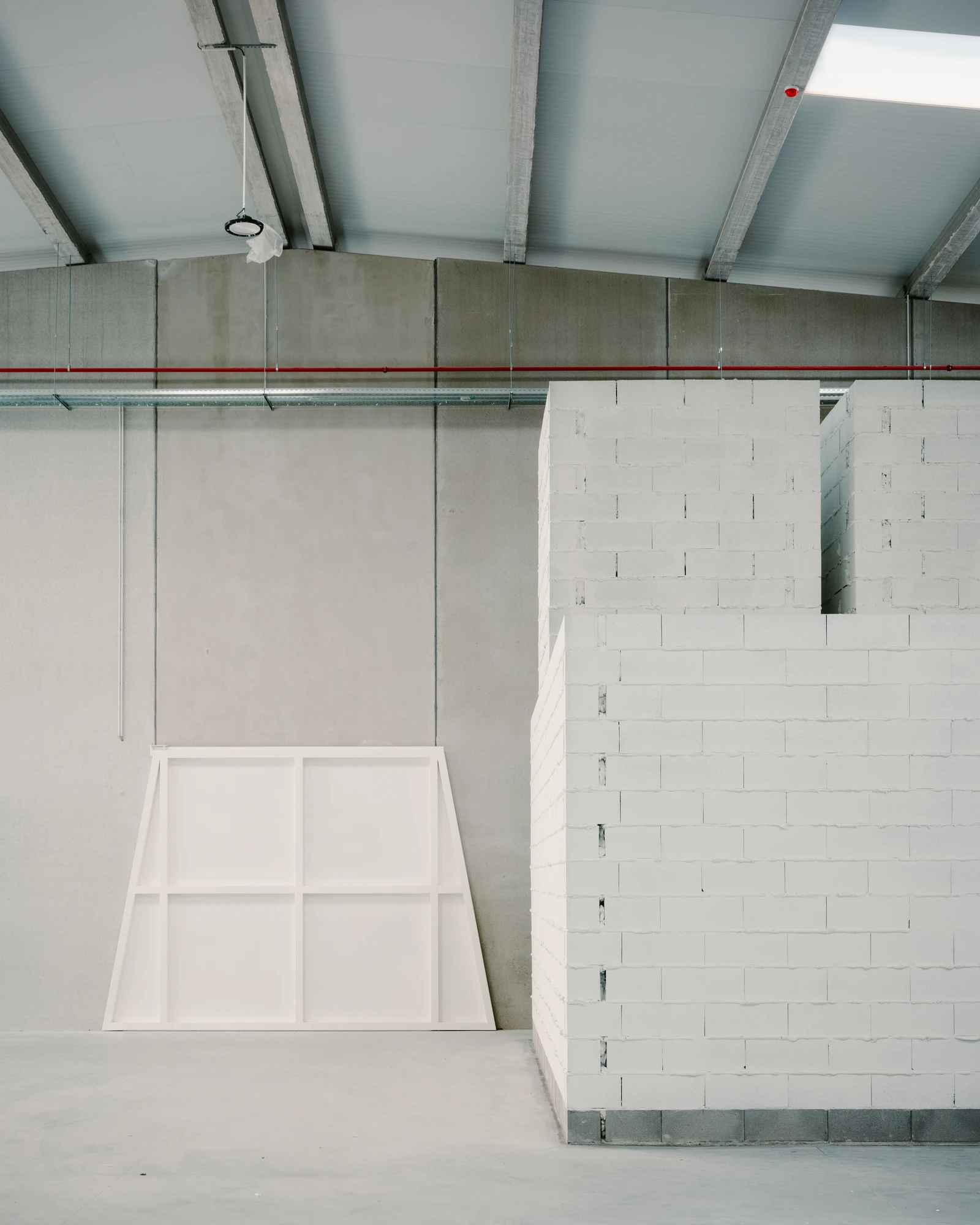 Corcet Headquarters by Nuno M. Sousa, photographed by Federico Farinatti, Francisco Ascensão — A working hall sits under a pitched metal roof on concrete beams, a red service pipe crossing the wall and pale block volumes rising at right.