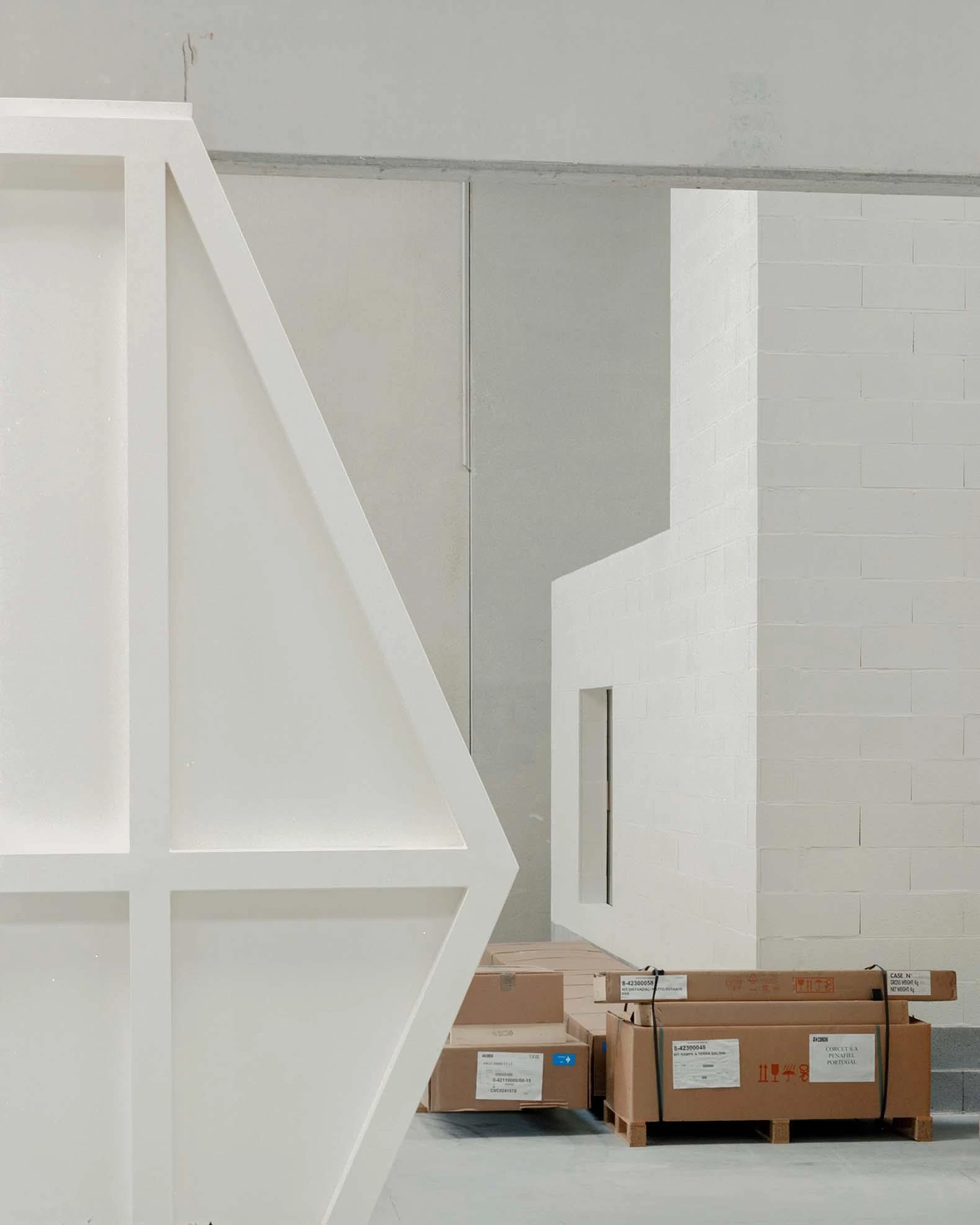 Corcet Headquarters by Nuno M. Sousa, photographed by Federico Farinatti, Francisco Ascensão — A white triangular frame stands beside a pale block volume, with shipping crates stamped for the Penafiel client stacked on the floor behind.