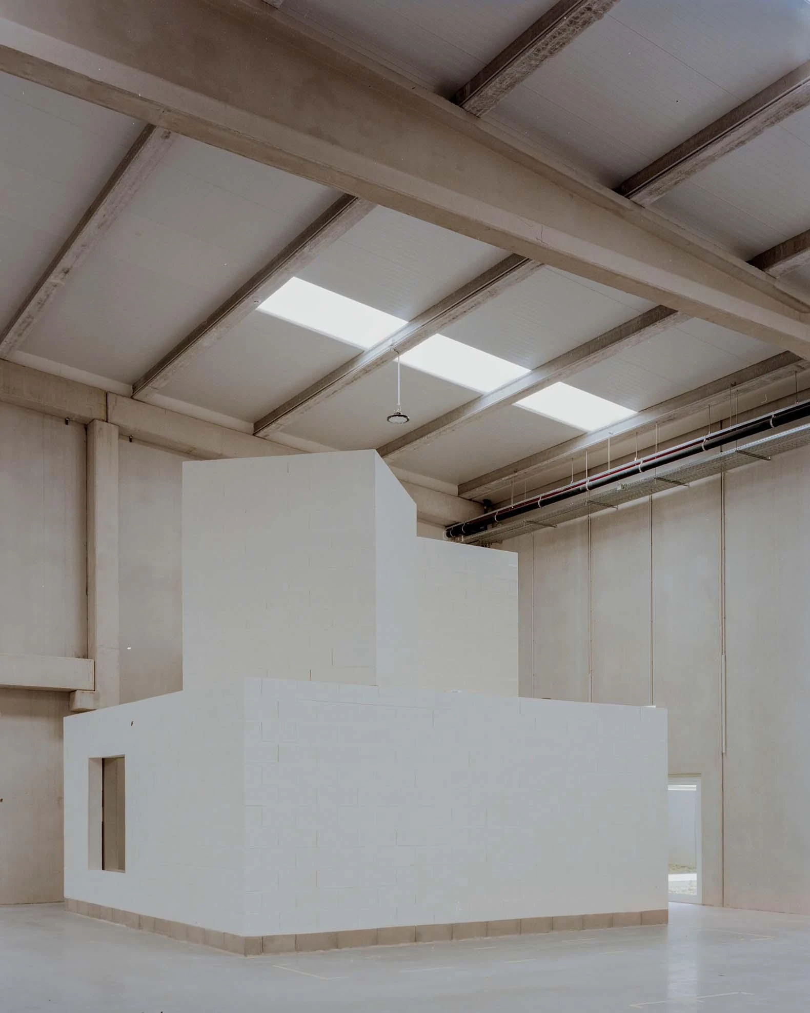 Corcet Headquarters by Nuno M. Sousa, photographed by Federico Farinatti, Francisco Ascensão — A skylit hall steps up in pale block volumes beneath an exposed pitched roof, the concrete floor stretching away toward a far opening.