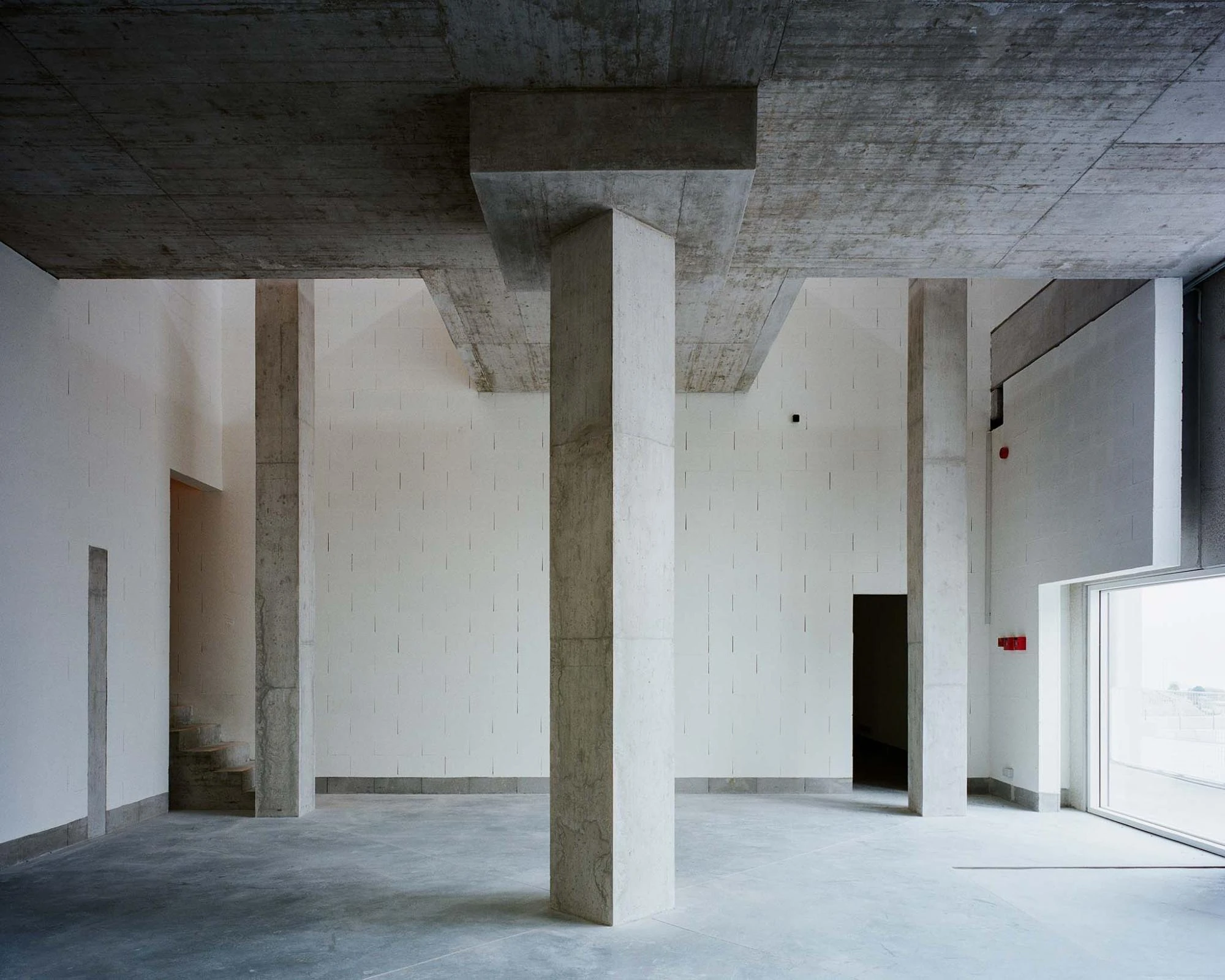 Corcet Headquarters by Nuno M. Sousa, photographed by Federico Farinatti, Francisco Ascensão — The double-height lobby gathers four columns under heavy concrete beams, a short flight of steps at left and full-height glazing spilling light at right.